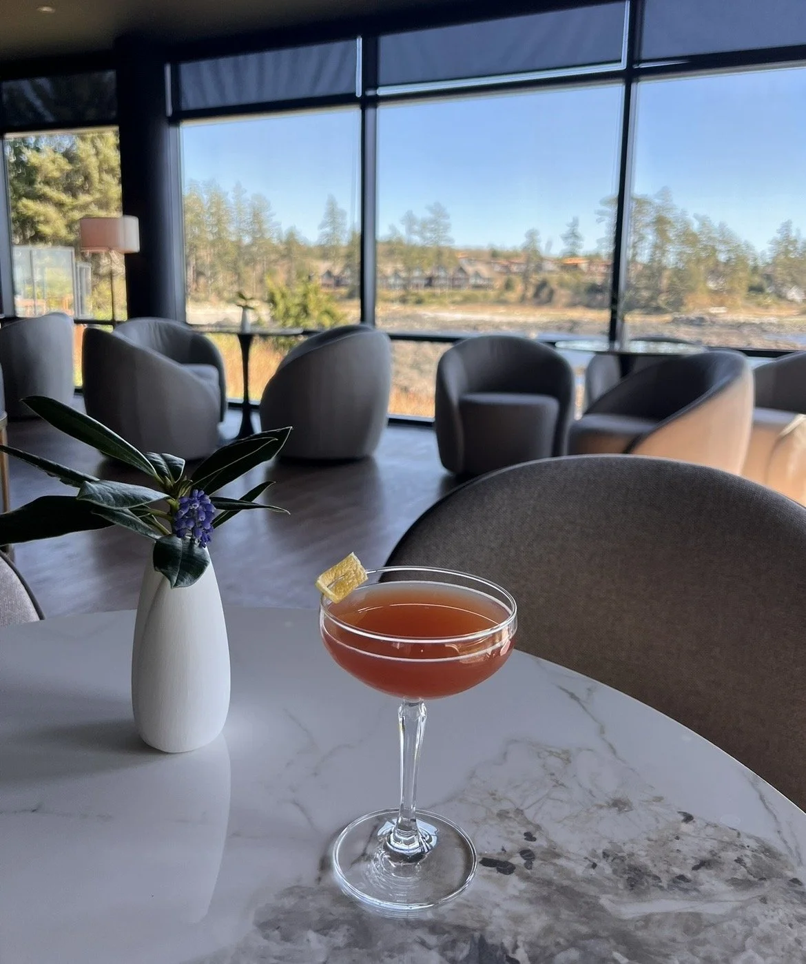 A lounge or bar area with large windows overlooking a scenic outdoor view, a white vase with green leaves and purple flowers on a white marble table. Commercial Design from Spirit design studio - Vancouver island interior decorator