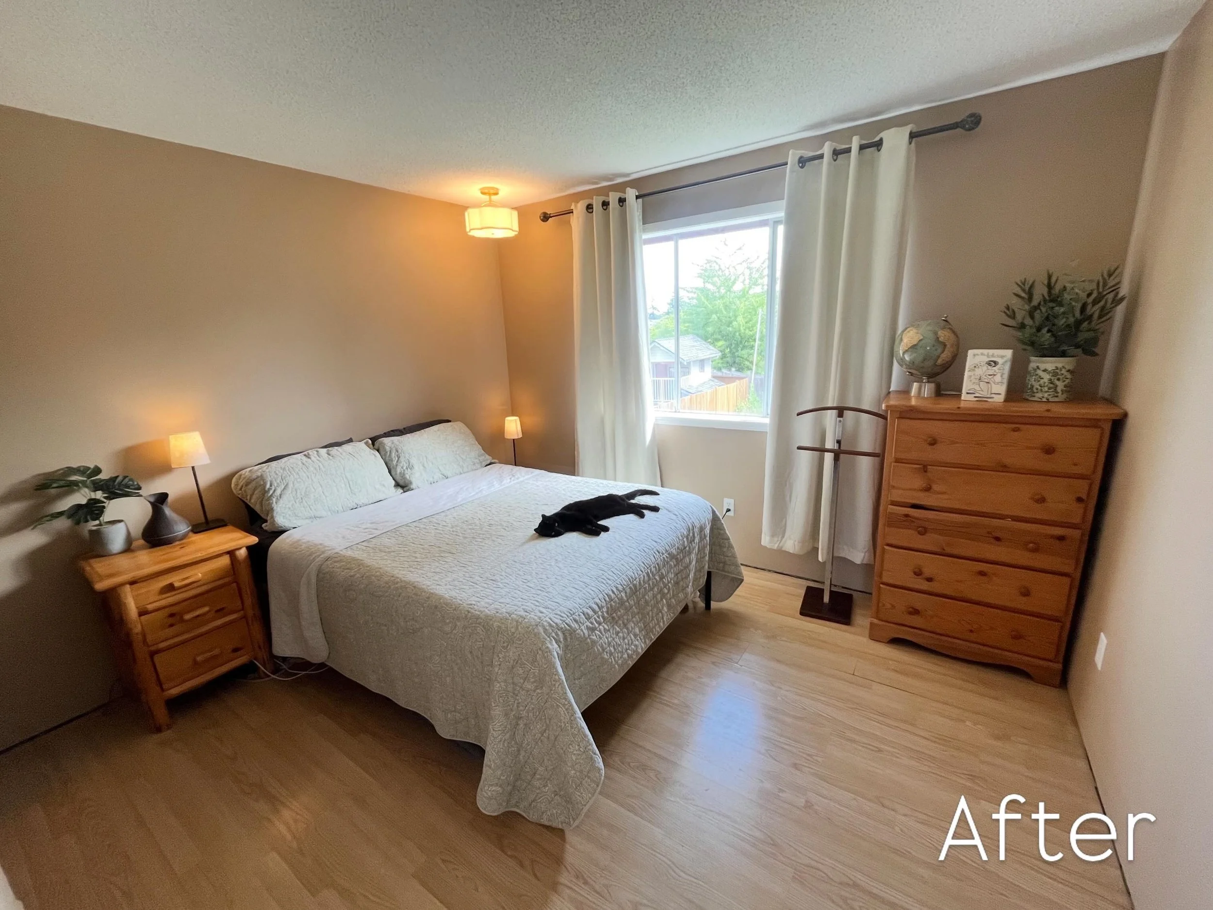 A tidy bedroom with a bed, wooden nightstand, dresser, and window with white curtains, sunlight outside, and a black cat lying on the bed. North Cowichan/ Duncan area , Spirit Design Studio, designed by Skylar Holman