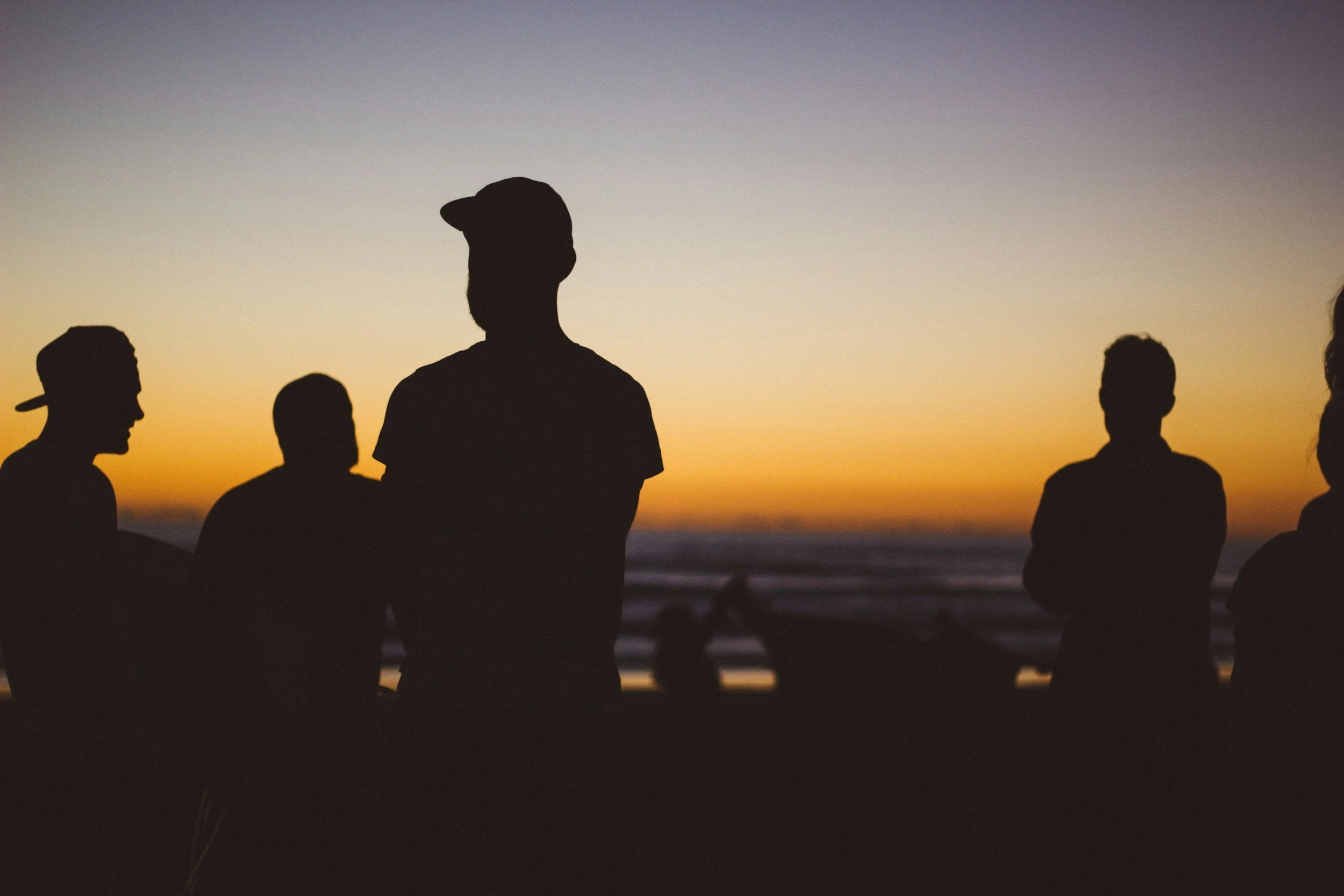 Silhouettes of five people standing together outdoors at sunset by the beach.