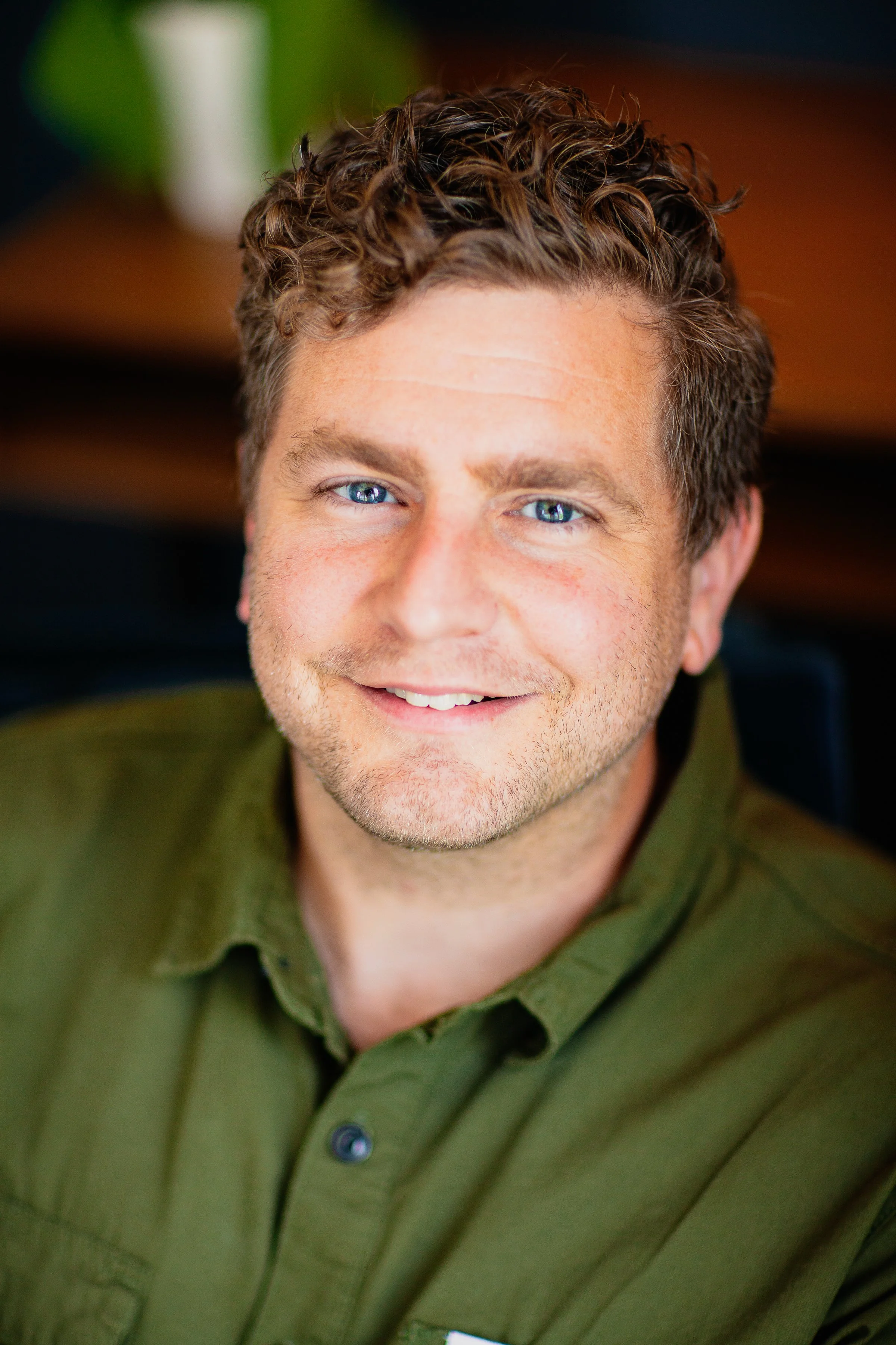 A young man with curly brown hair and blue eyes smiling, wearing a green shirt.