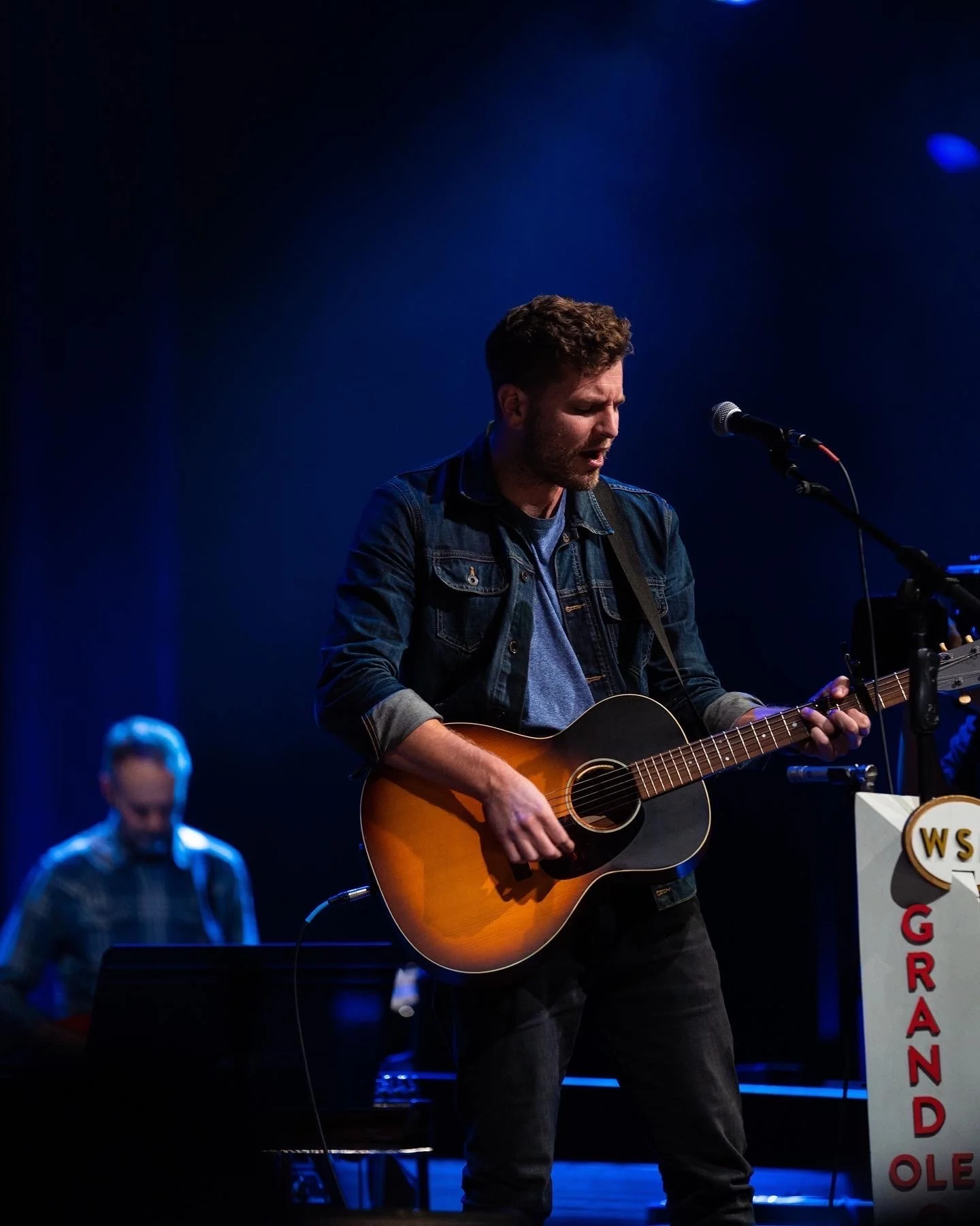 A man playing an acoustic guitar singing into a microphone during a performance on stage, with a woman in the background playing a keyboard.