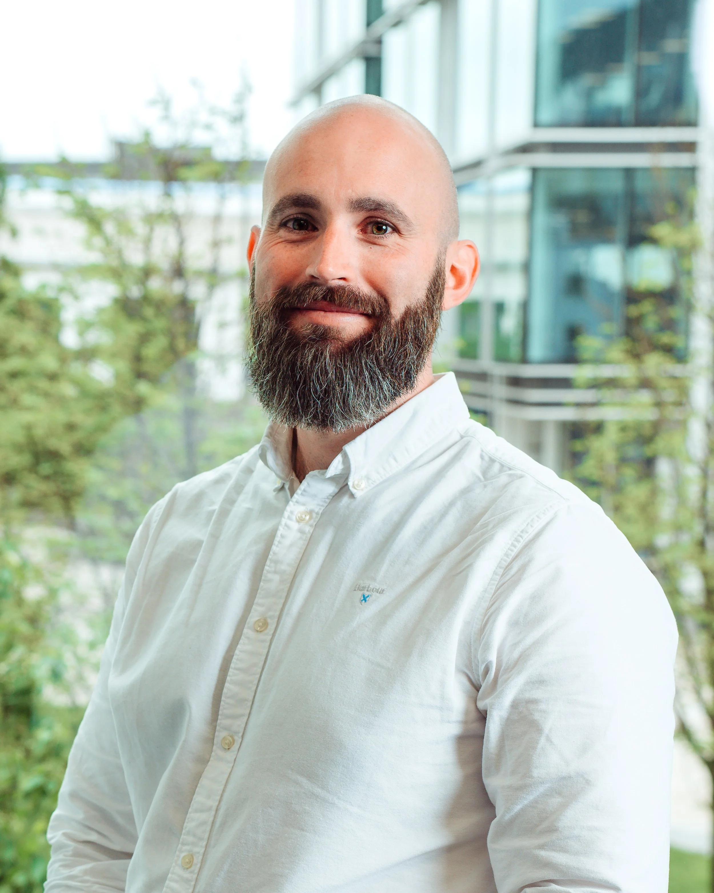 A man with a beard and a shaved head, wearing a white button-up shirt, smiling slightly, standing indoors with large windows and a modern building structure visible in the background.