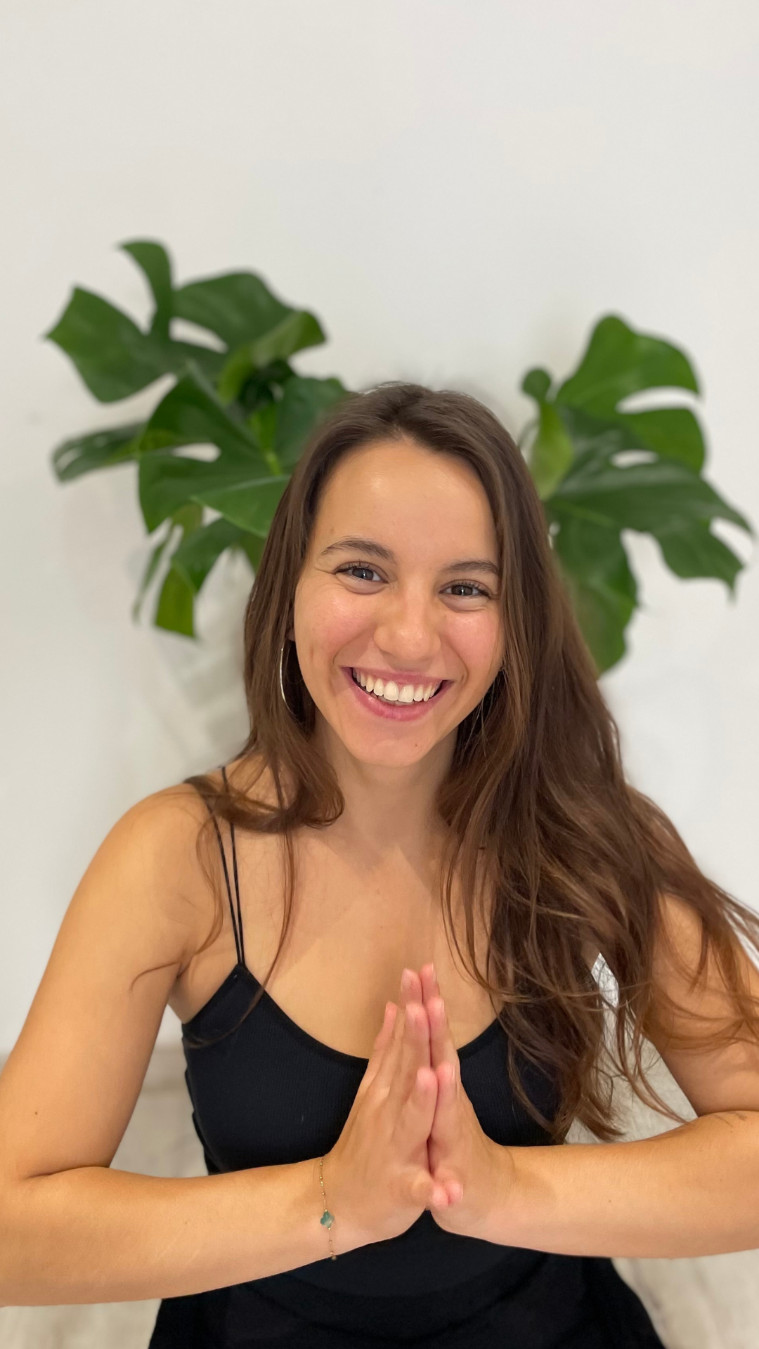 Une femme souriante, yeux brillants, en position de prière, assise devant une plante verte.