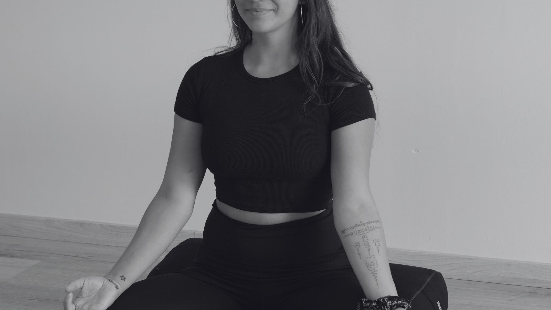 Une femme assise en position de méditation, portant un t-shirt noir cropped et des pantalons sombres, assise sur un tapis par terre contre un mur blanc, avec des tatouages sur ses bras.