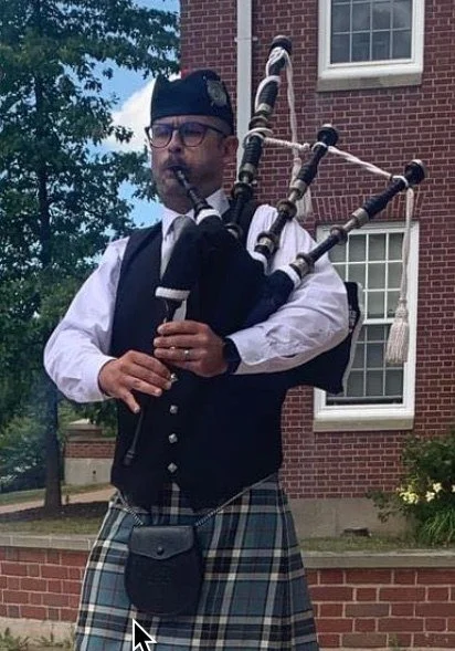 A man dressed in traditional Scottish attire playing bagpipes outside in front of a brick building.