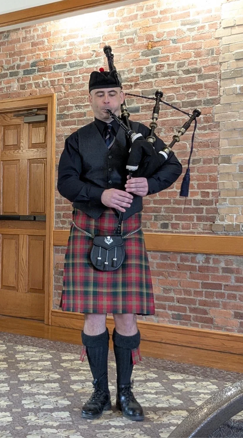 A man dressed in traditional Scottish attire, including a kilt, playing bagpipes indoors near a brick wall.