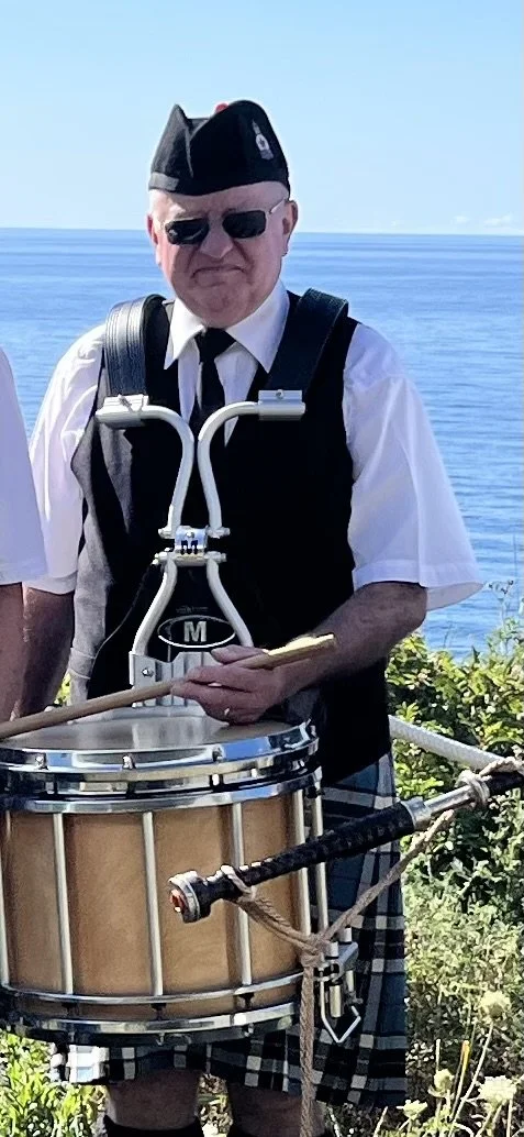 A man wearing a black beret, sunglasses, white shirt, and vest, playing a snare drum outdoors near the ocean, with greenery and blue sky in the background.