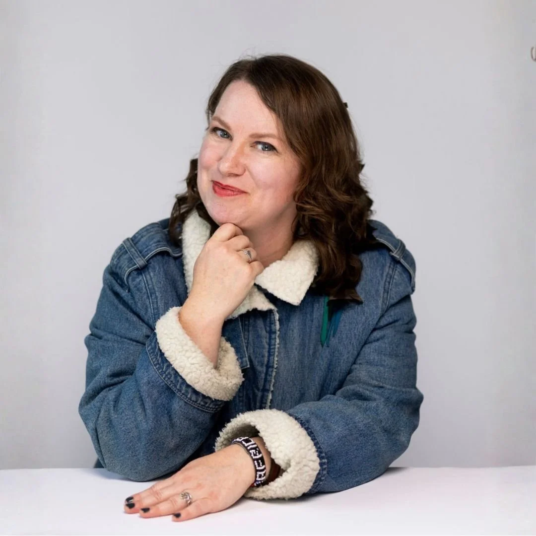 A woman with shoulder-length wavy brown hair and fair skin, sitting at a table with a plain light gray background, wearing a denim jacket with a cream fuzzy lining, resting her chin on her left hand, smiling at the camera.