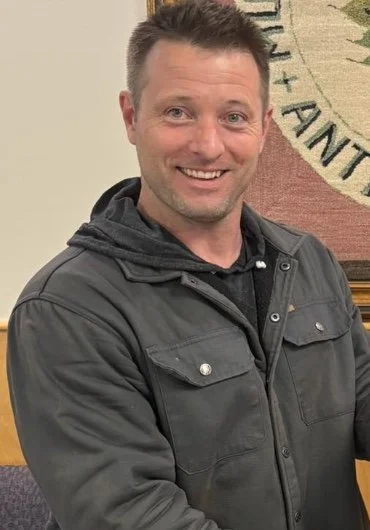Smiling man with short brown hair, wearing a dark jacket, standing indoors in front of a wall and a large emblem or banner.
