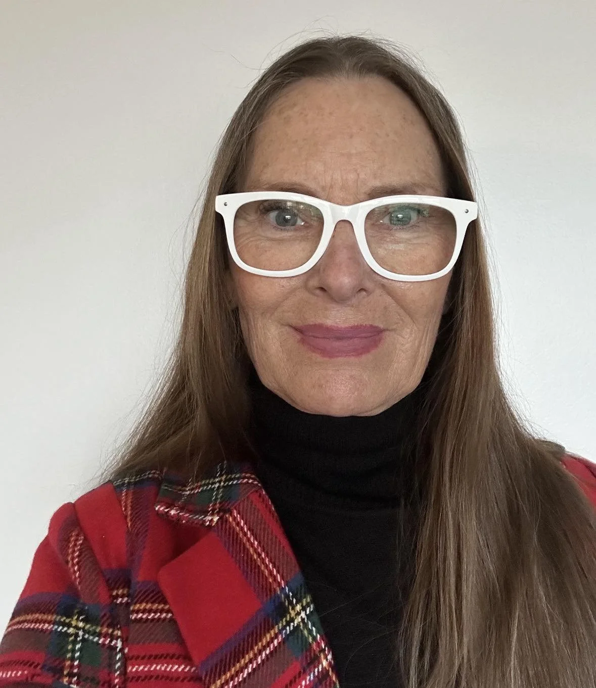 A woman with long brown hair, wearing white glasses, a black turtleneck, and a red plaid blazer, standing against a plain white wall.