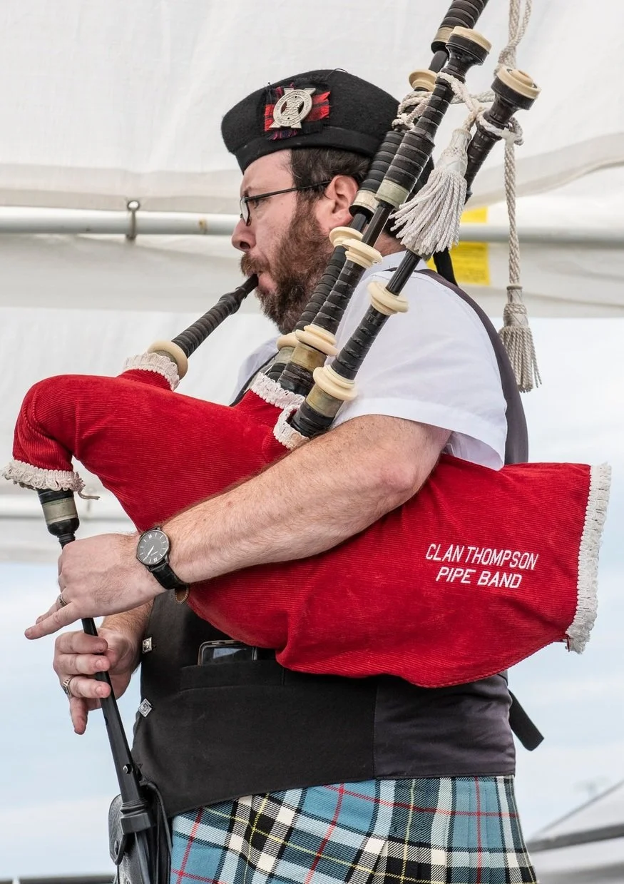 A man in traditional Scottish attire playing bagpipes, with a kilt, vest, and a Glengarry cap, under a tent.