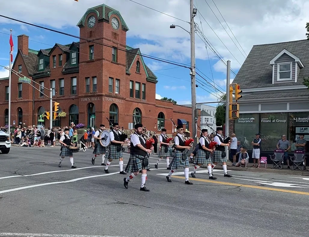 A parade with participants dressed in traditional Scottish kilts and playing bagpipes moves across a crosswalk in front of a town hall building. Onlookers are gathered on the sidewalk, some sitting and some standing, while some are taking photos. The town hall is a large brick building with a clock tower, and the sky is partly cloudy.