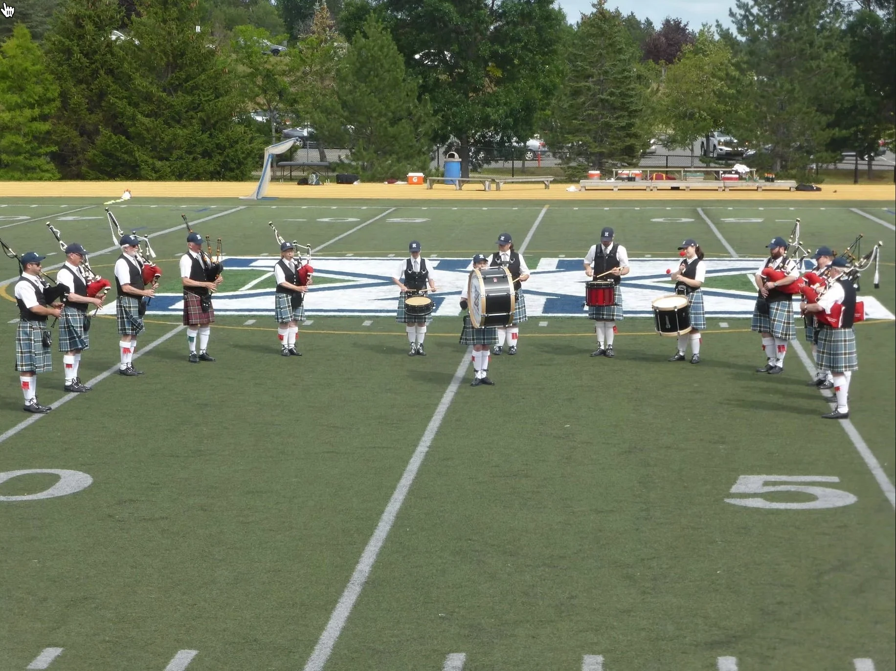 A group of band members dressed in traditional Scottish attire playing bagpipes and drums on a football field with a large logo, trees, and benches in the background.
