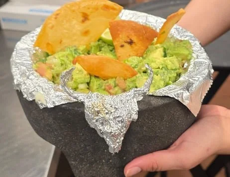 A hand holding a stone bowl with guacamole topped with tortilla chips and a slice of cheese.