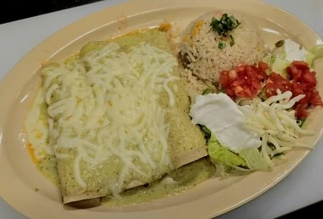 A plate of Mexican food with a burrito covered in green sauce, shredded cheese, a serving of rice with cilantro, and a small salad with lettuce, sliced tomatoes, sour cream, and shredded cheese.