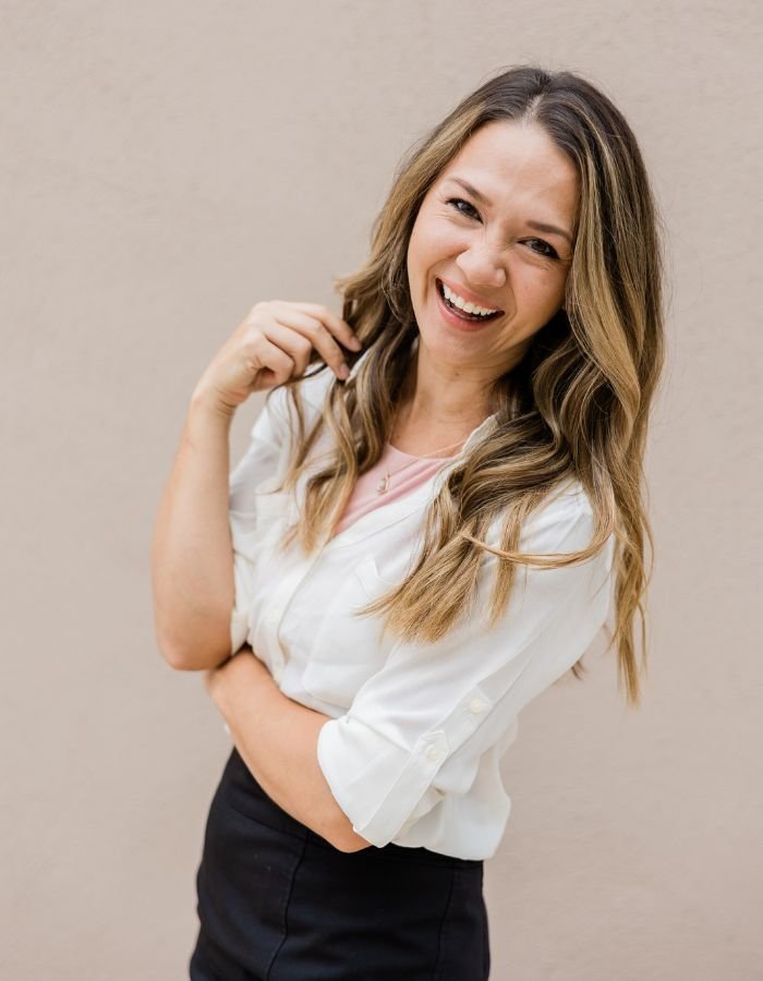 Happy woman with long, wavy, blonde hair wearing a white blouse and black skirt, standing against a beige wall.