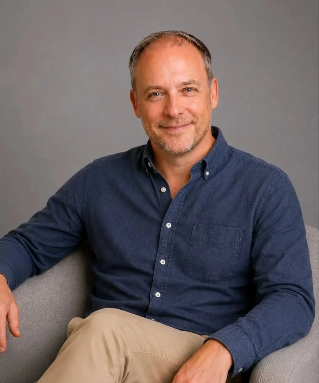 Charles Taylor,  therapist in Redondo Beach, California, with short, light brown hair and blue eyes, wearing a navy blue button-down shirt and beige pants, sitting on a gray sofa against a gray background with a slight smile.