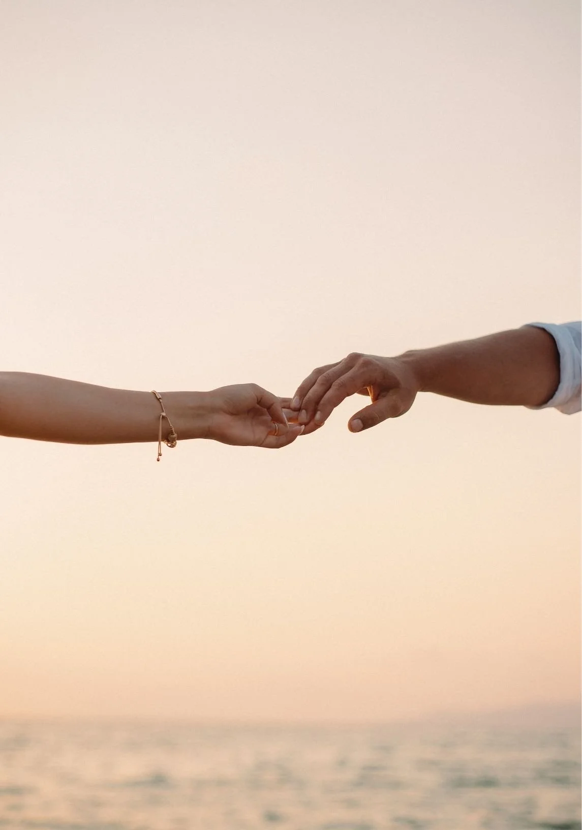 Two people reaching out and holding each other's fingers against a soft peach-colored sky.