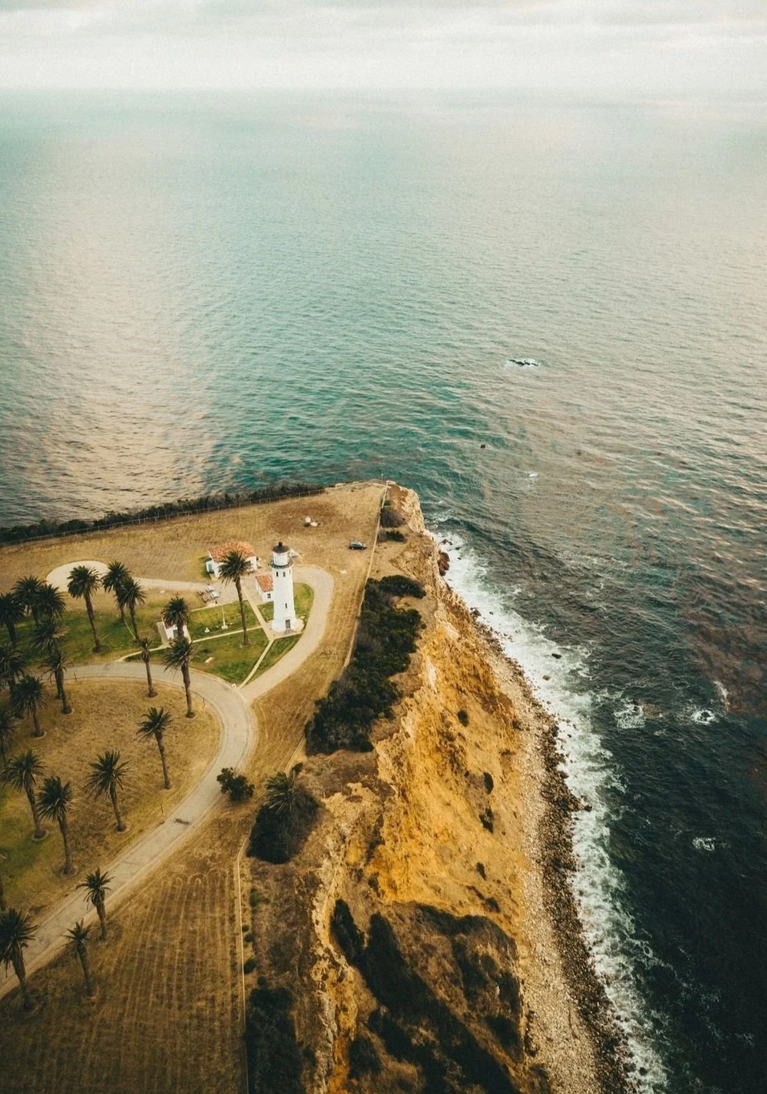 Aerial view of a lighthouse on a rocky coast with a grassy area and palm trees, overlooking the ocean with waves hitting the shoreline.