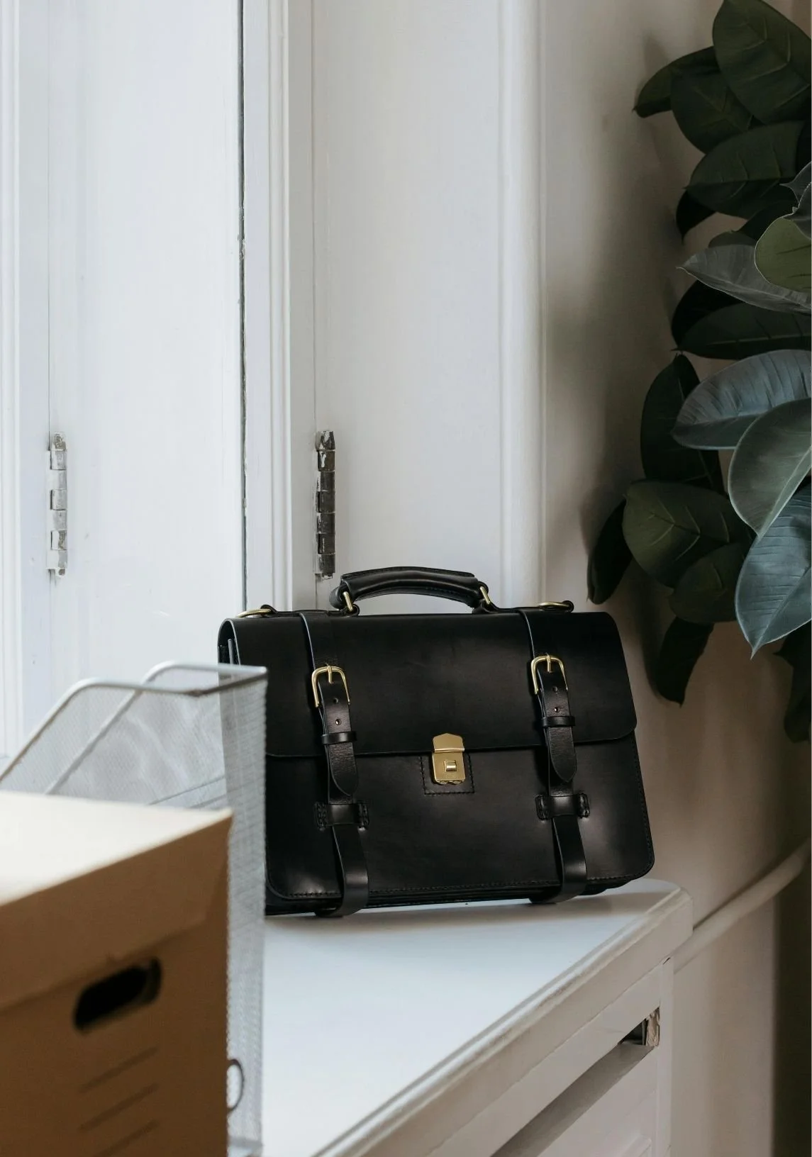 A black leather briefcase with gold hardware sitting on a white cabinet, with part of a cardboard box and a white wire basket visible in the foreground. A green leafy plant is on the right side, and a white door with a metal hinge is in the background.