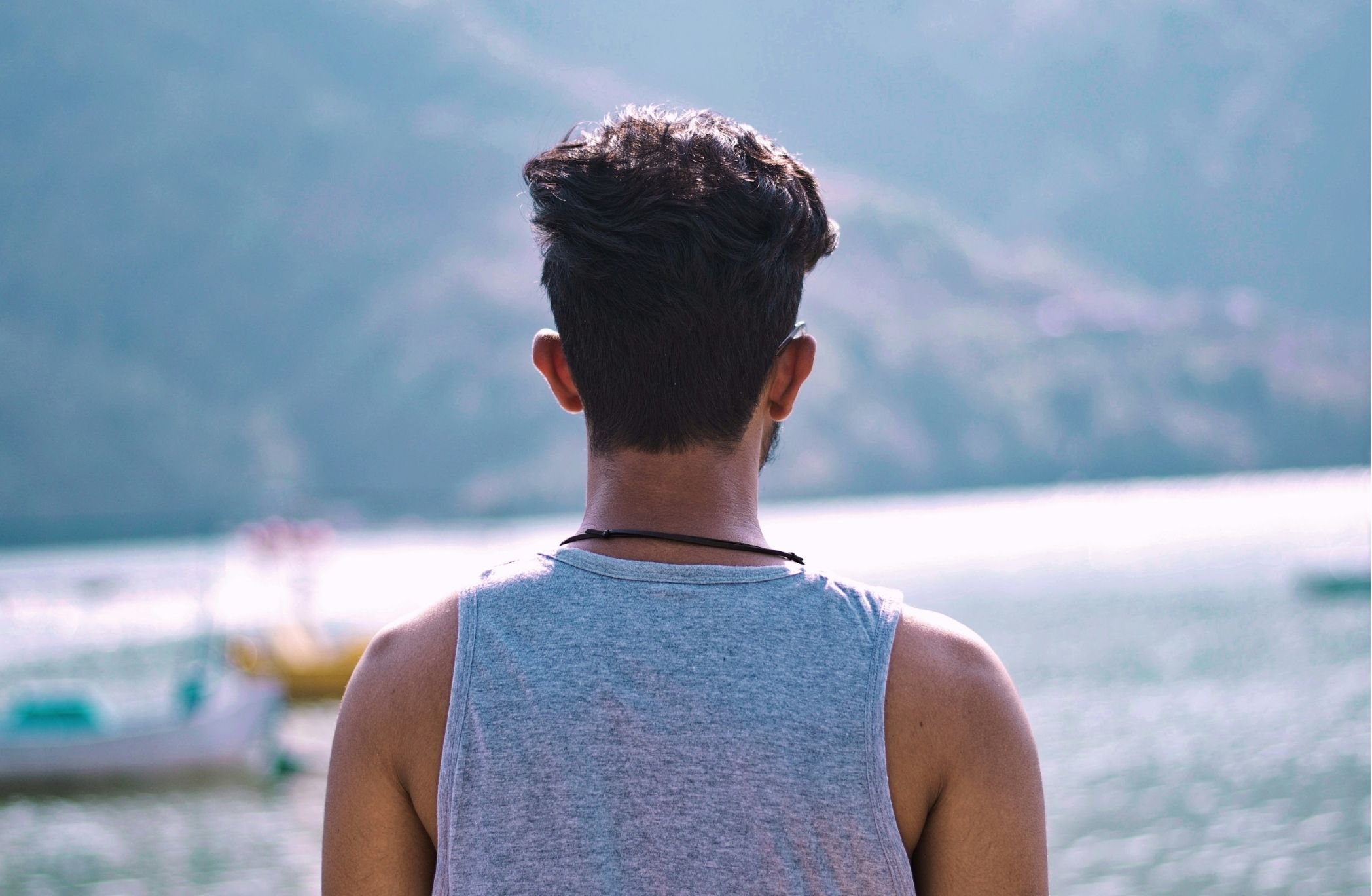 Back view of a young man with short dark hair wearing a sleeveless gray shirt, looking out at a body of water with blurred boats and distant shoreline.