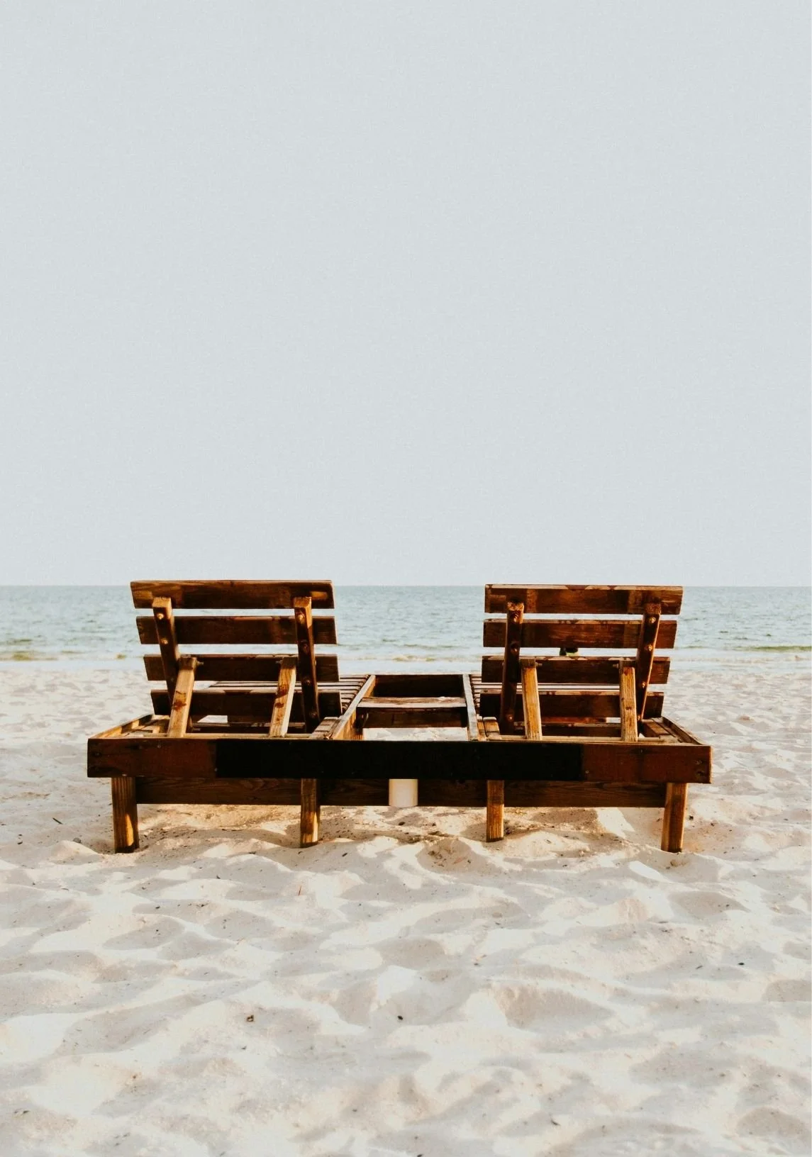 Two wooden lounge chairs on a sandy beach facing the ocean, positioned side by side with a small table in between, under a clear sky.