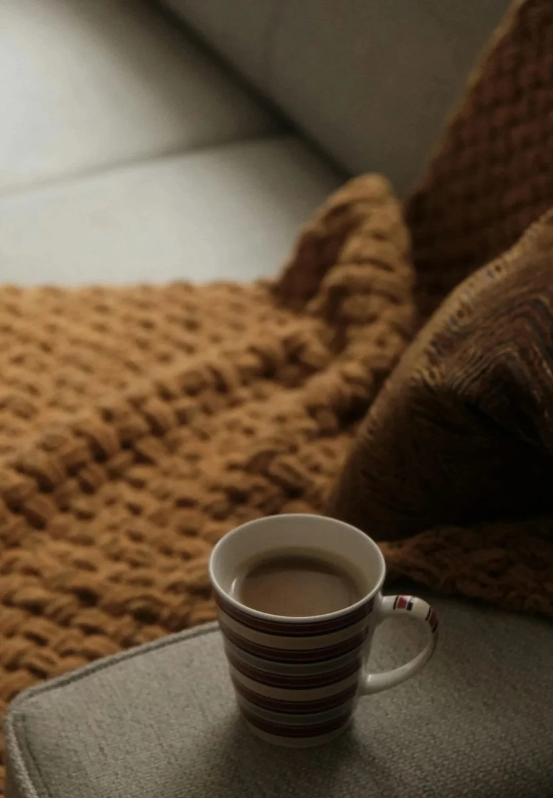 A striped coffee mug filled with coffee on a beige surface, with a brown textured blanket and a white wall in the background.