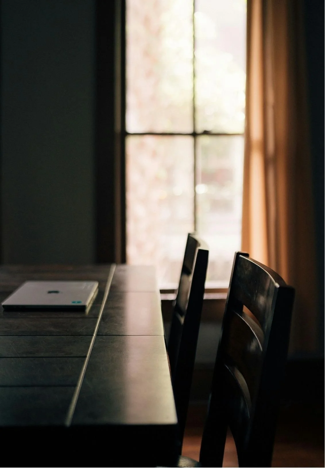 A dimly lit dining room with wooden chairs lined up along a dark wooden table. In the background, a window with light curtains allows soft sunlight to enter, creating a cozy, peaceful atmosphere.