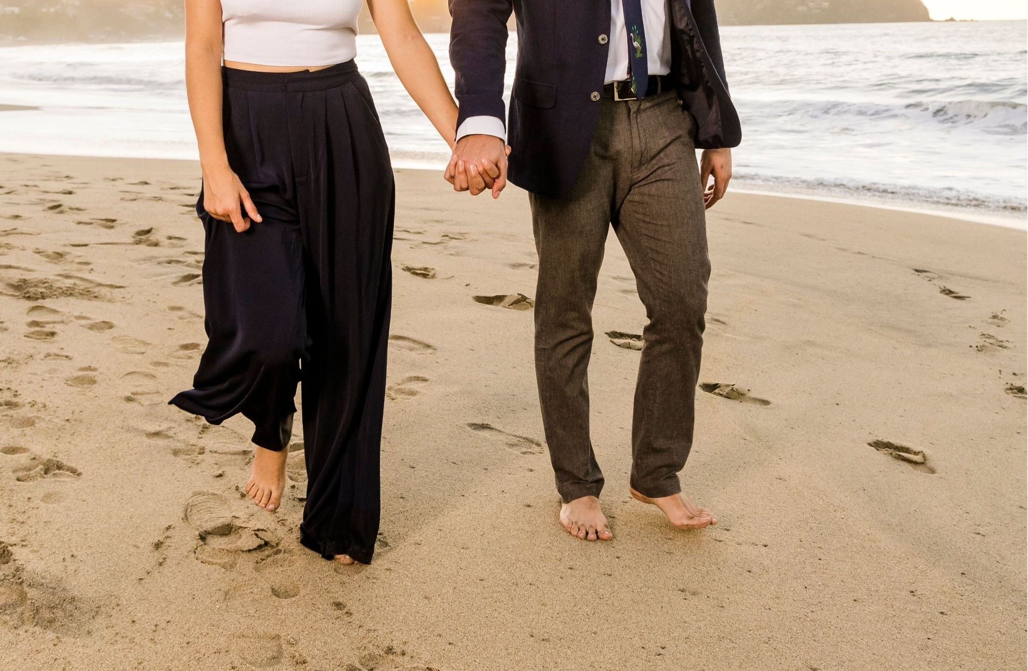 A couple holding hands and walking barefoot on the beach, visible from the waist down. The woman is wearing loose black pants and a white crop top, while the man is in brown trousers, a dark blazer, and a shirt with a tie featuring a crane design. They are leaving footprints in the sand as they walk near the ocean.