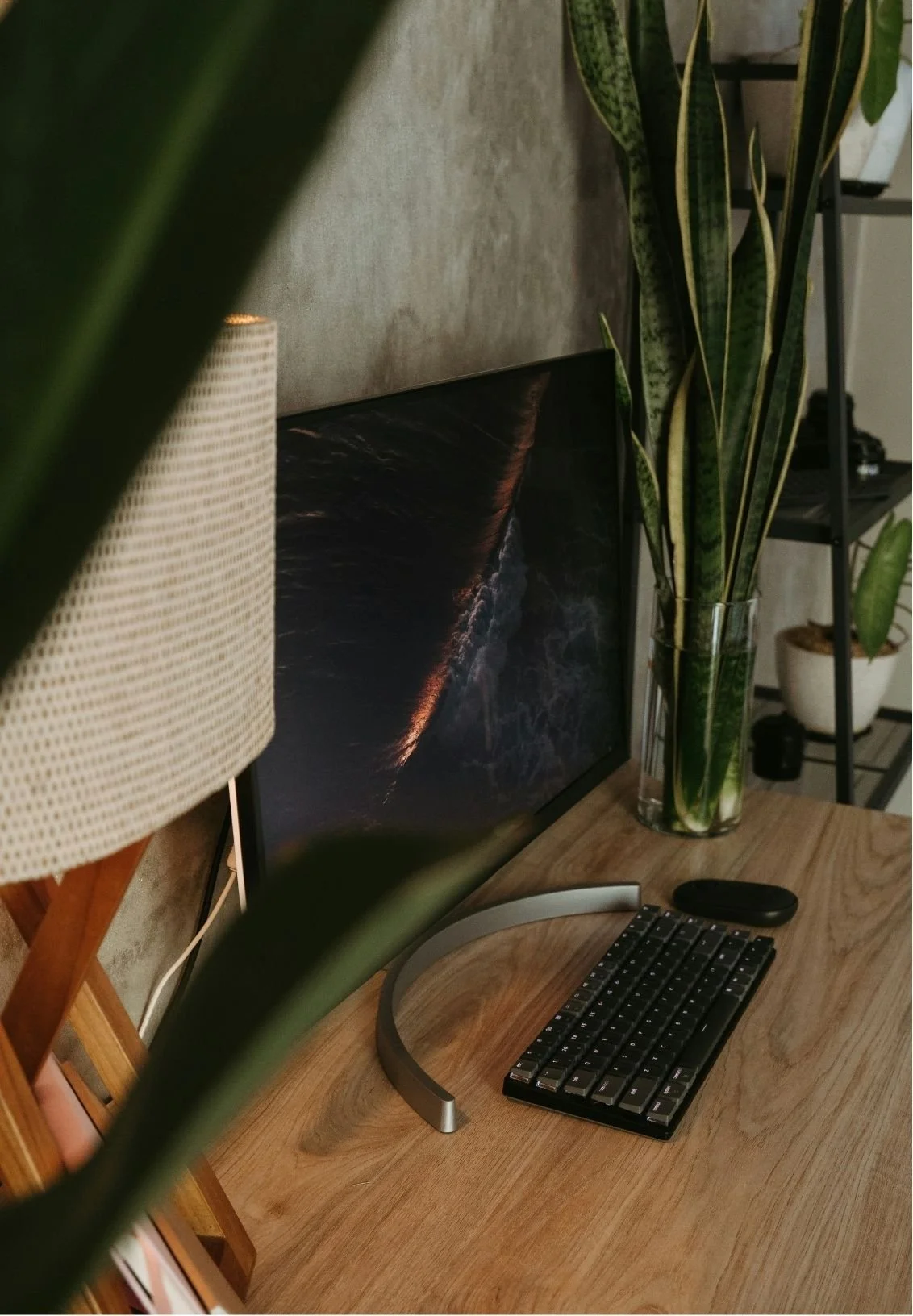 A computer desk with a large monitor displaying a dark ocean scene, a wireless keyboard, a mouse, potted plants, and a lamp in a cozy, well-decorated home office.