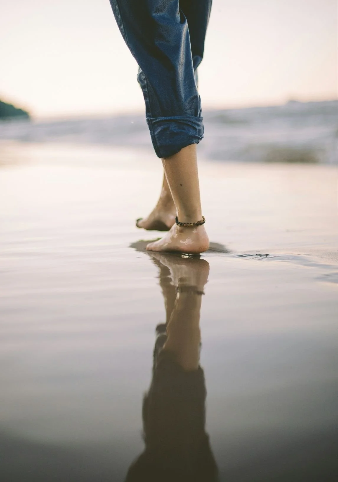 Person walking barefoot on a wet sandy beach, rolled-up jeans, anklet on left ankle, sunset or sunrise in the background.