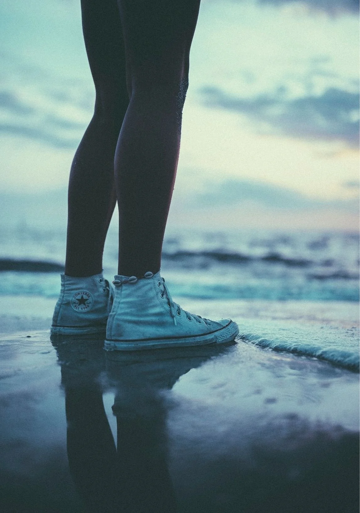 Close-up of a person wearing white Converse sneakers standing on the wet sand at the beach with the ocean in the background during dusk.