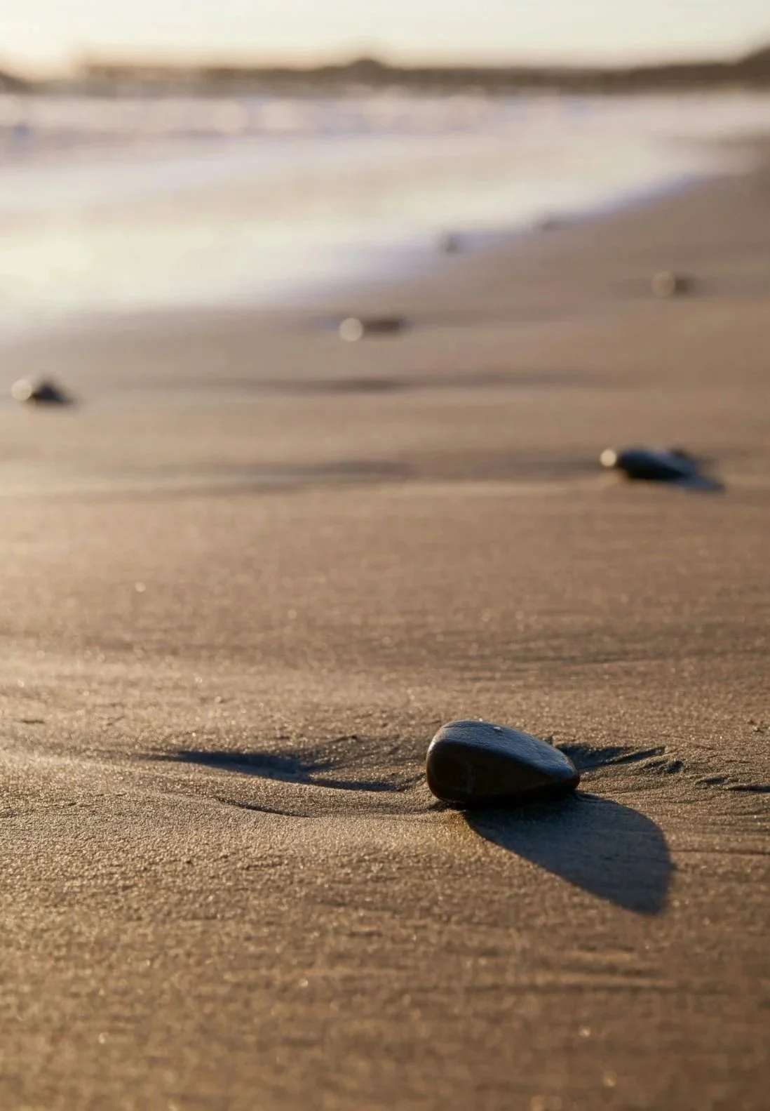 Close-up of a smooth black stone on sandy beach with other stones scattered in the background, ocean waves and horizon in the distance during sunset.