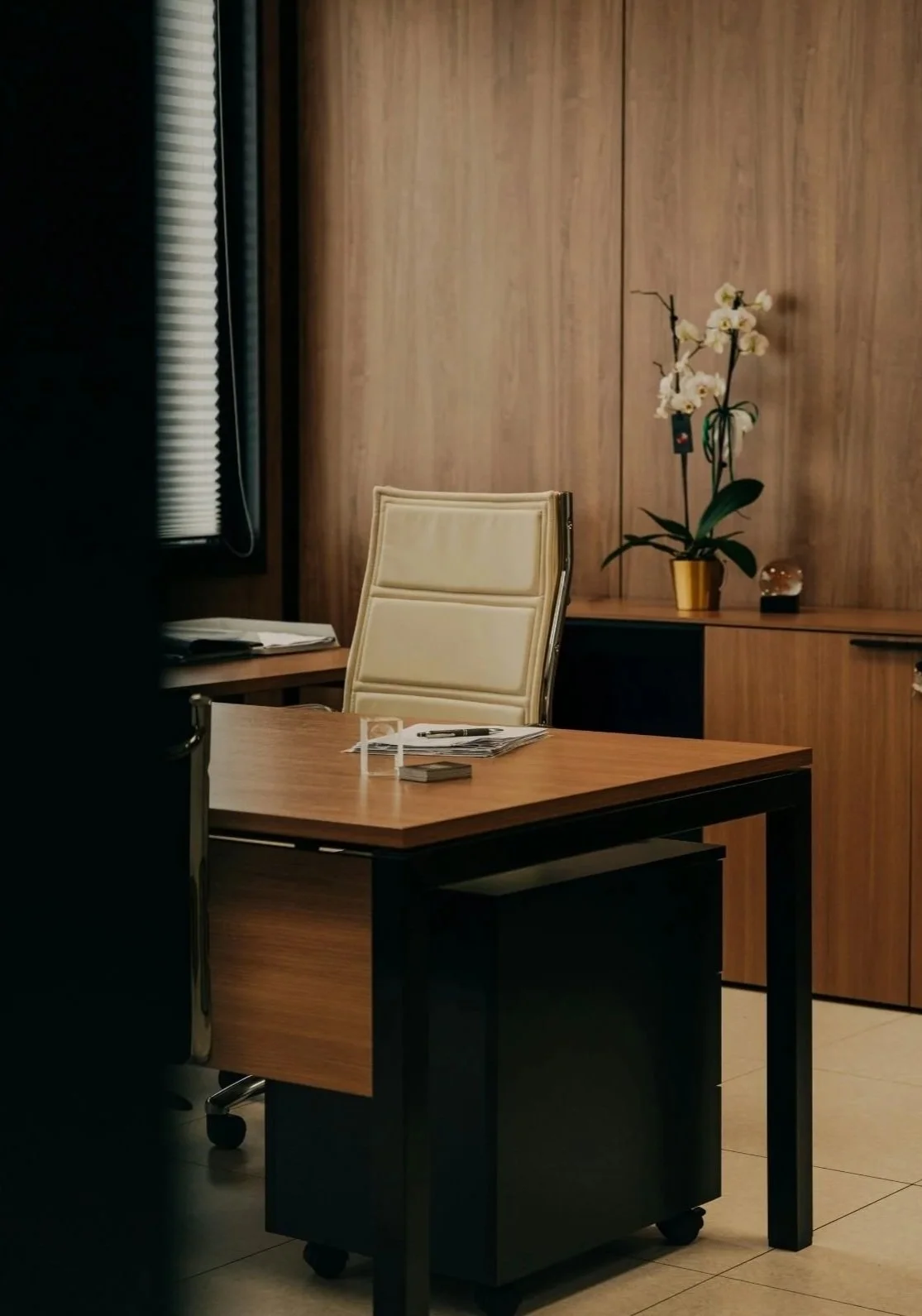 An empty office with a wooden desk, a cream-colored chair, a potted orchid on a cabinet, and a window with closed blinds.