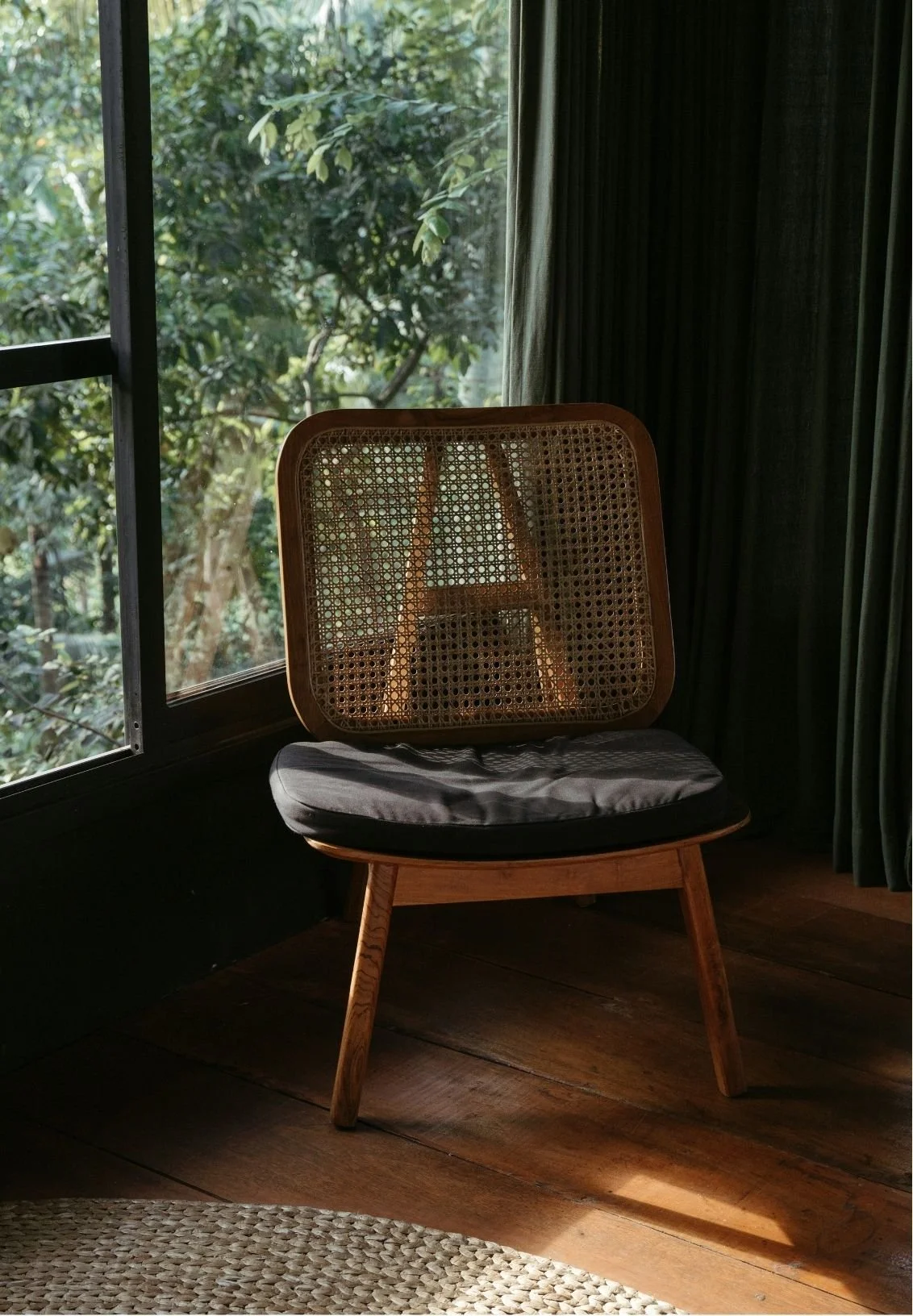 A wooden chair with a woven cane backrest and a black cushioned seat, positioned near a window with lush green foliage outside, in a dimly lit room with dark curtains and wooden floor.