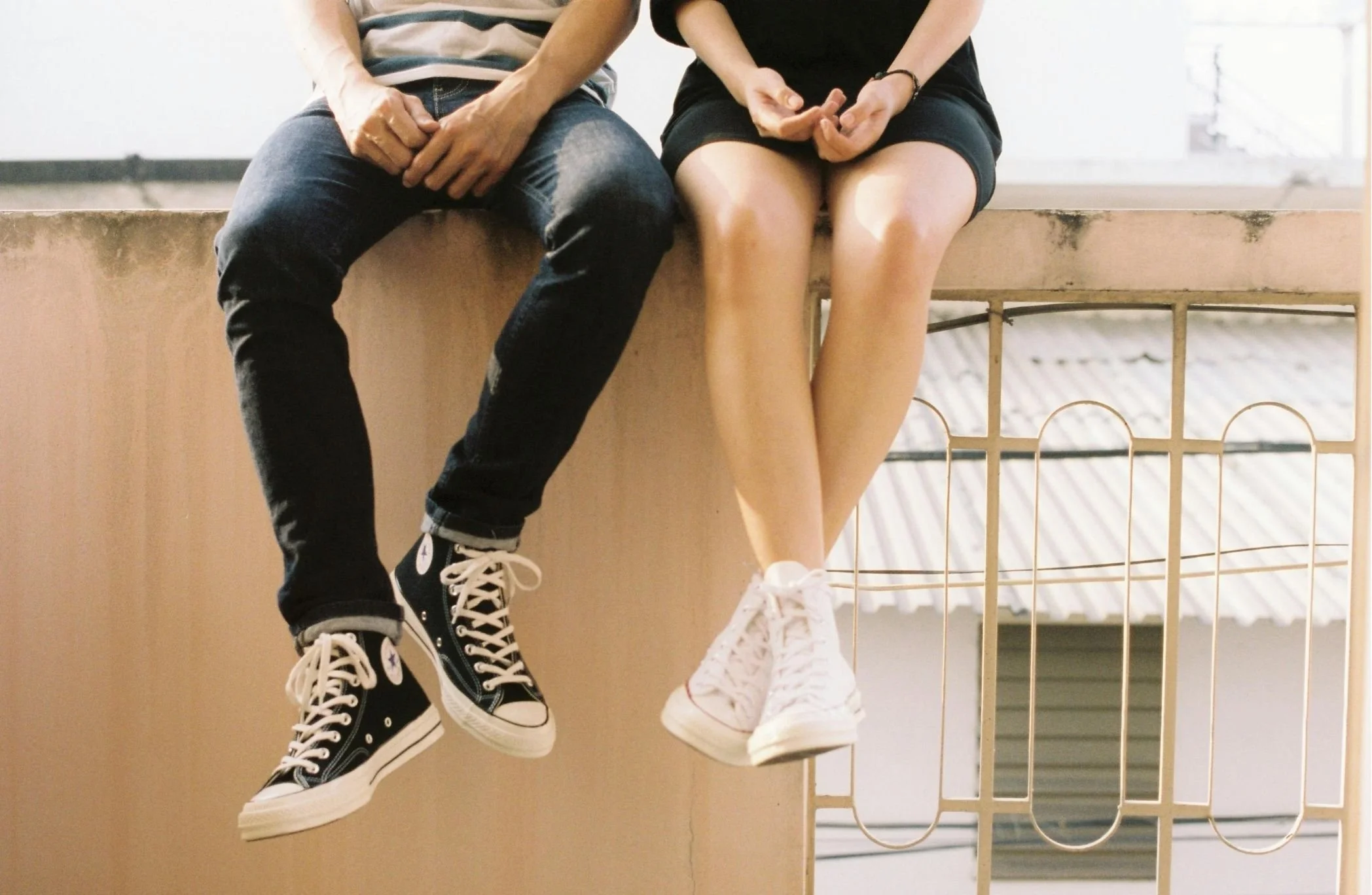 Two people sitting side by side on a windowsill, holding hands with their legs hanging down. They are wearing casual clothing, high-top sneakers, and shorts.