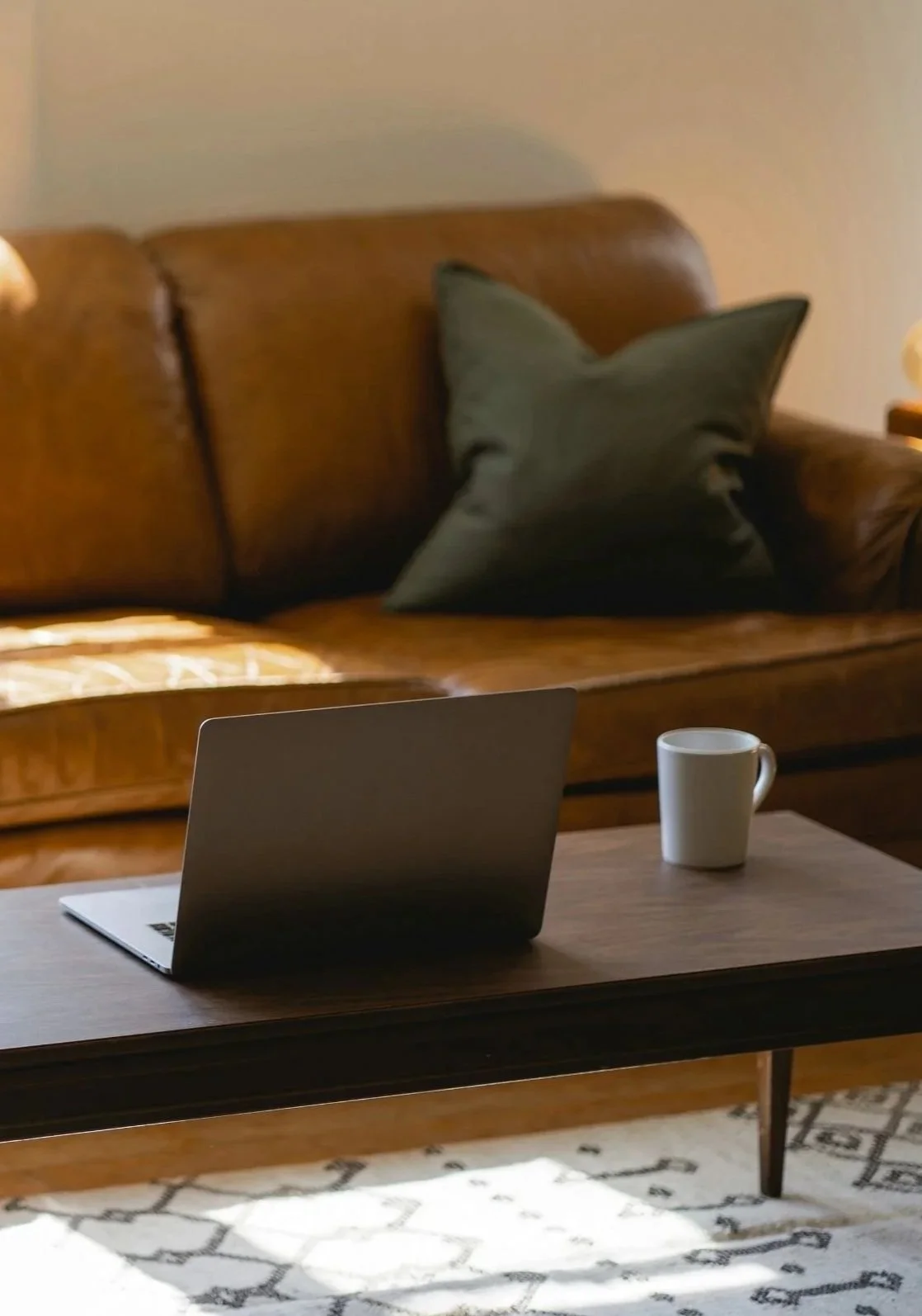 Living room with a brown leather sofa, dark green pillow, wooden coffee table with a laptop and a white mug, and a patterned area rug.