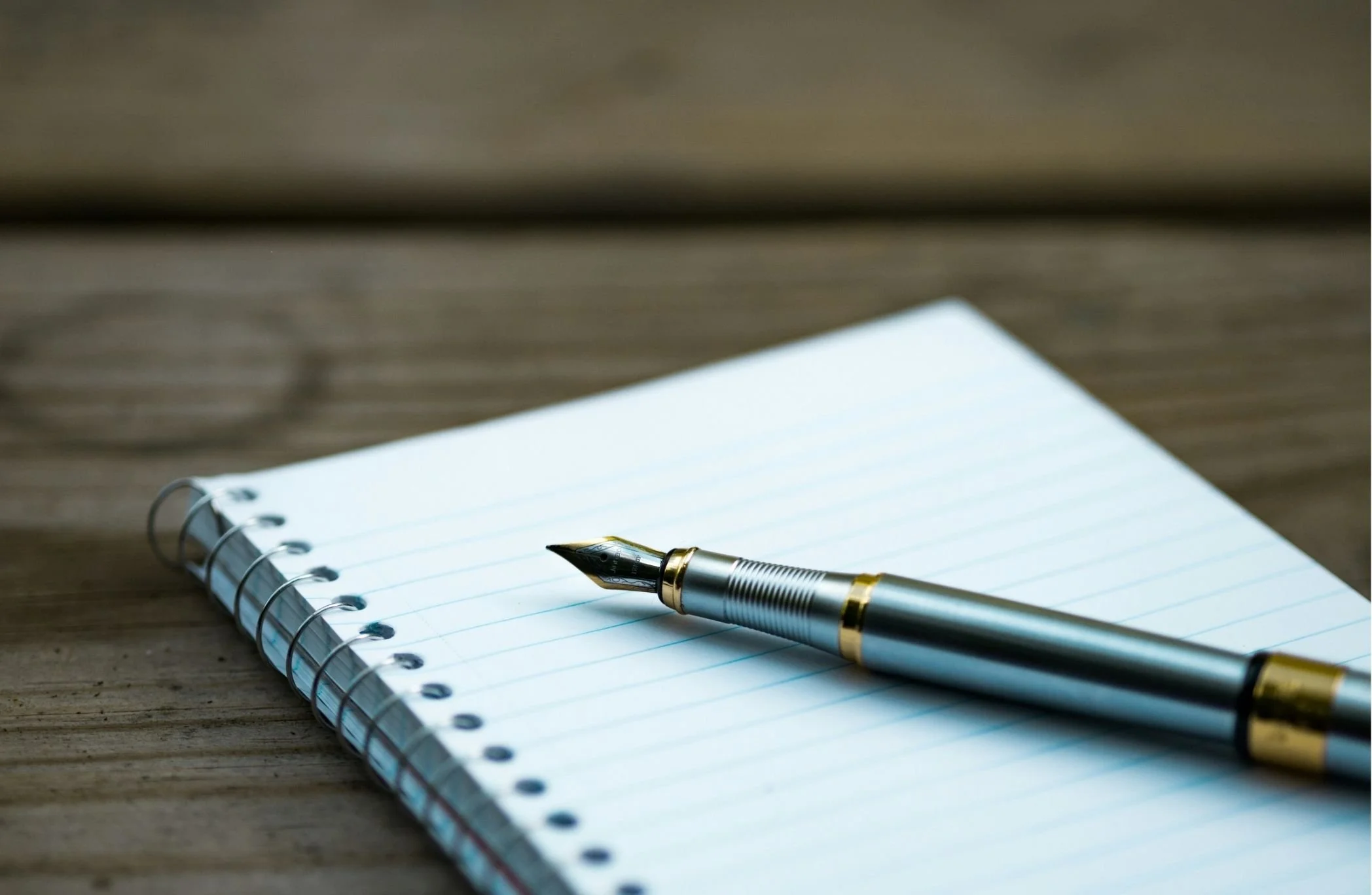 An open spiral notebook with blank white lined pages and a silver and gold fountain pen resting on it, placed on a wooden surface.