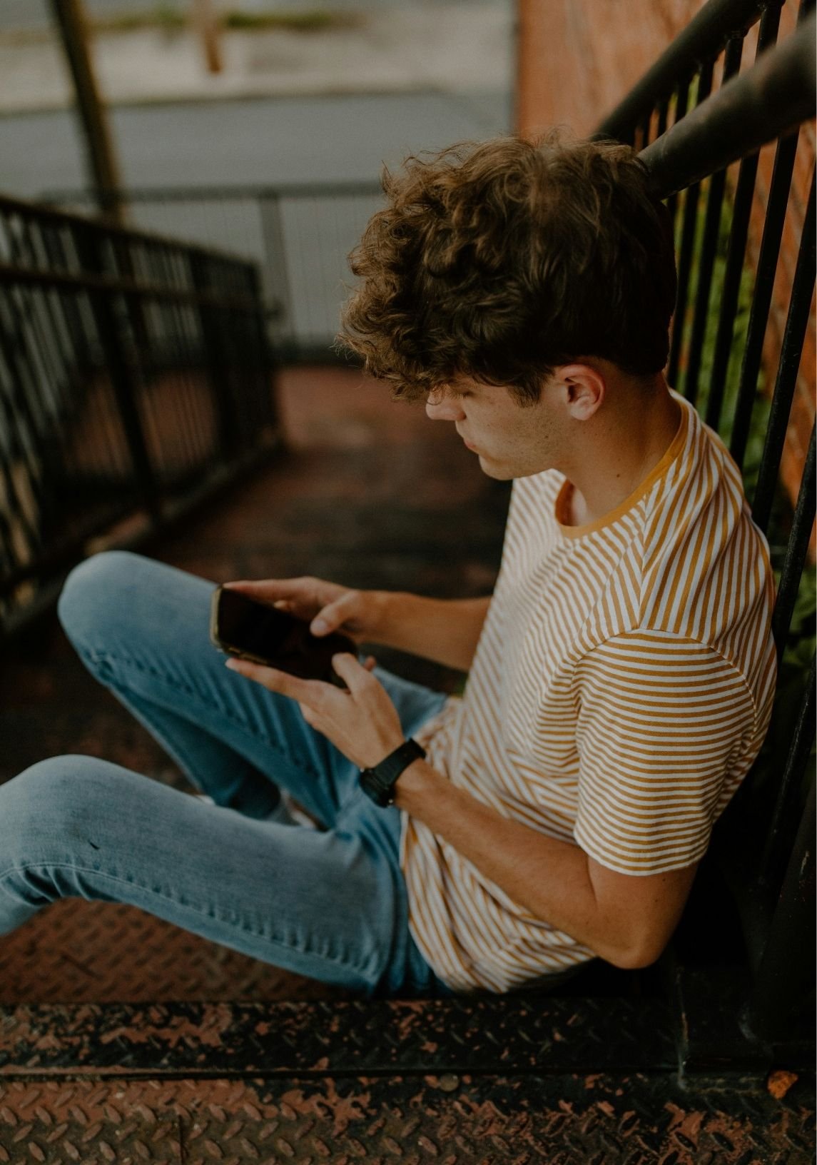 A young man with curly hair wearing a striped t-shirt, blue jeans, and a watch, sitting on a metal bench or staircase railing, looking at his smartphone in a dimly lit outdoor setting.