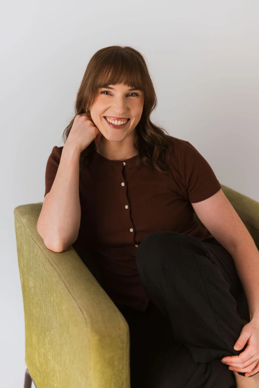 A woman with brown hair and bangs, smiling, sitting on a green sofa, wearing a brown short-sleeved top with buttons and black pants, resting her head on her left hand.