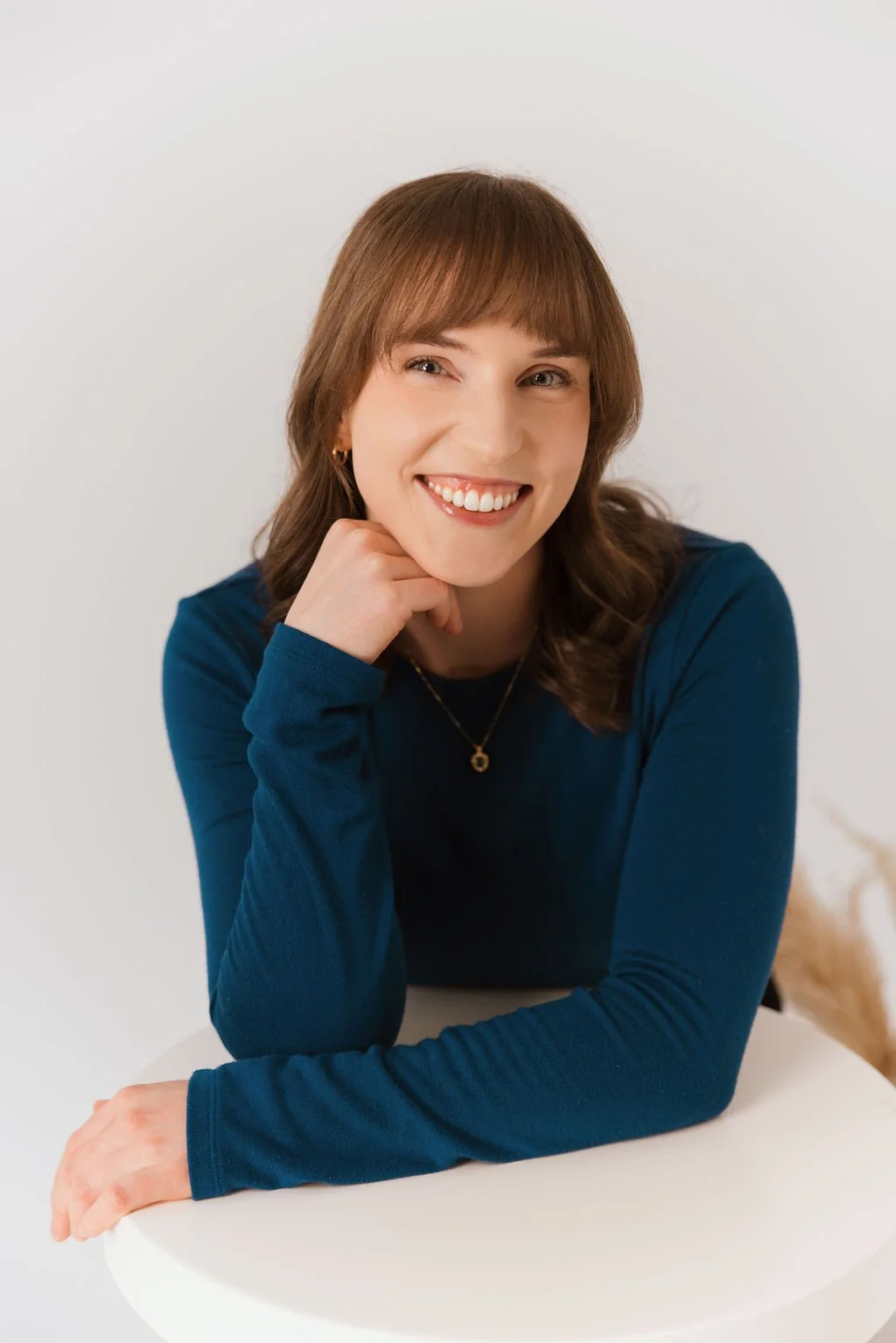 A woman with shoulder-length brown hair, wearing a blue long-sleeve shirt, sitting at a white table, smiling, resting her chin on her hand.
