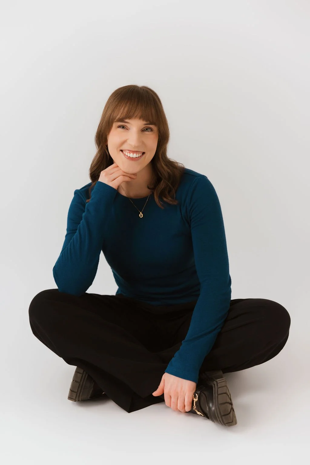 A young woman with brown hair and bangs, wearing a teal long-sleeve shirt, black pants, and black shoes, sitting cross-legged on a white background, smiling and resting her chin on her hand.