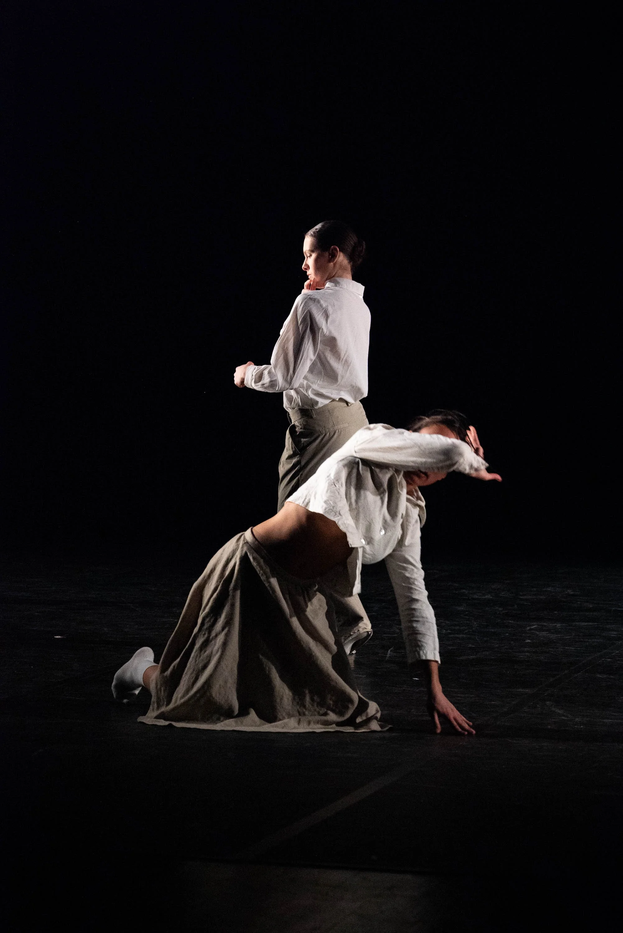 Two dancers perform on a black stage, with one standing and the other crouching or balancing on all fours, dressed in neutral-toned clothing.