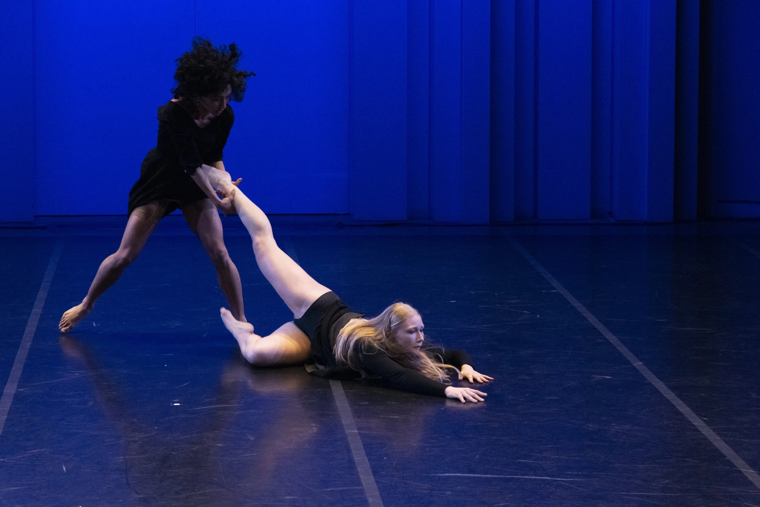 Two women performing a contemporary dance on a dark stage with blue curtains. One woman is lying on the floor on her stomach, while the other is standing and holding her arm, with a focus on movement and expression.