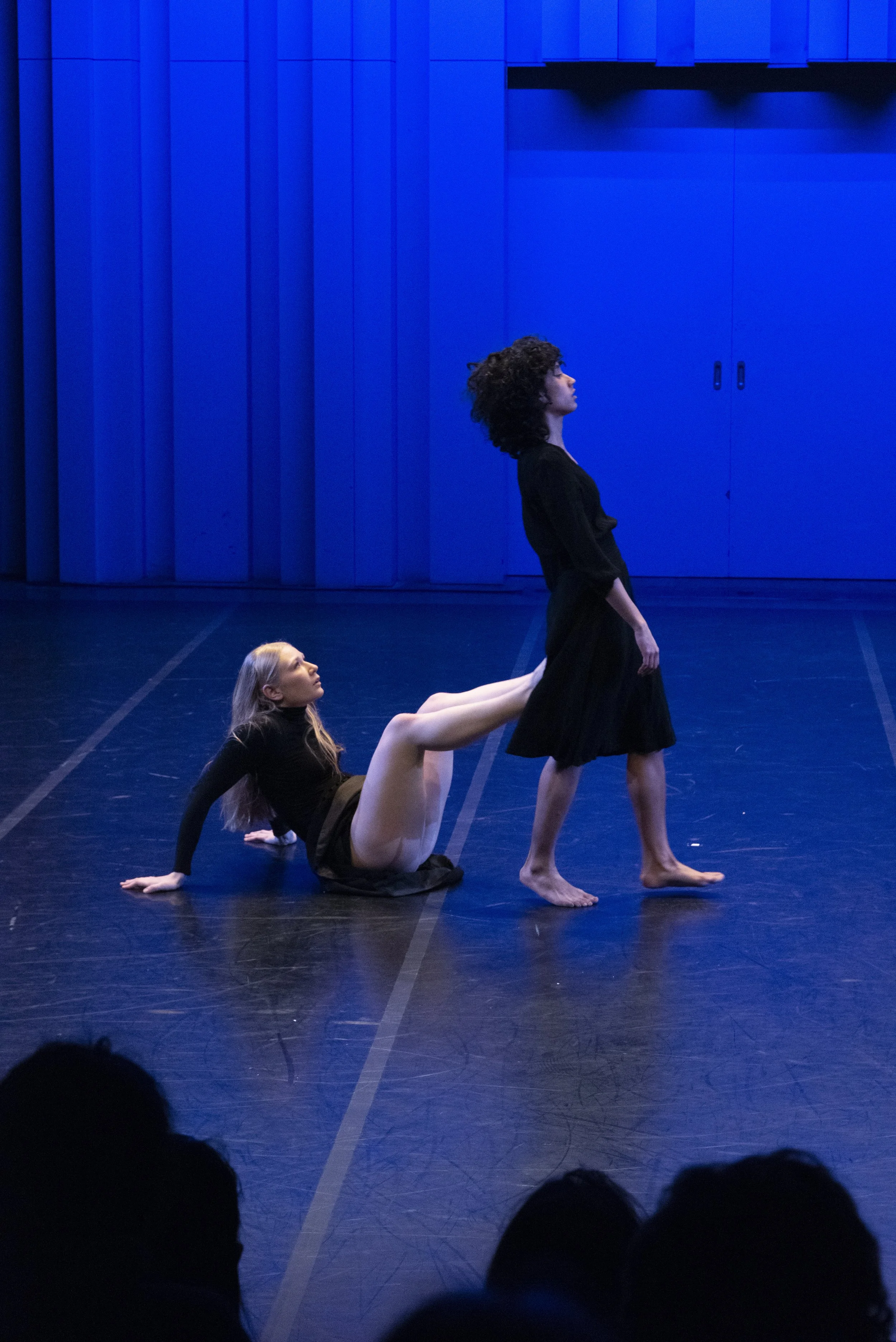 Three women performing a dance routine on stage with a blue backdrop. The woman in front stands upright, the woman in the middle is on her knees with her head tilted back, and the woman behind is on the floor supporting herself with her hands. They are dressed in black outfits.
