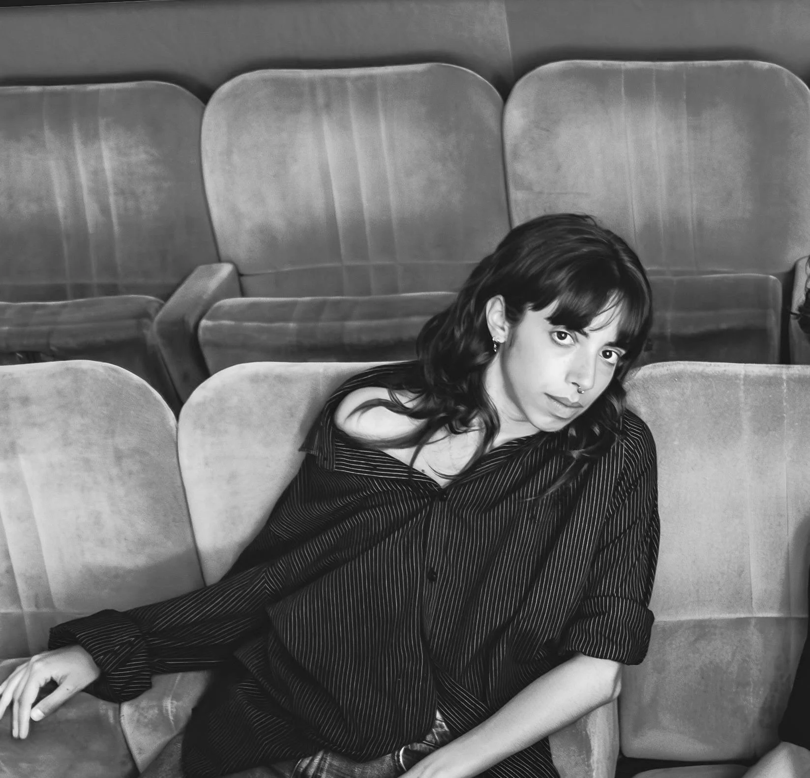 A young woman with dark hair and a septum piercing resting on a row of seats, wearing a dark, pinstriped shirt, in a setting with empty seats.