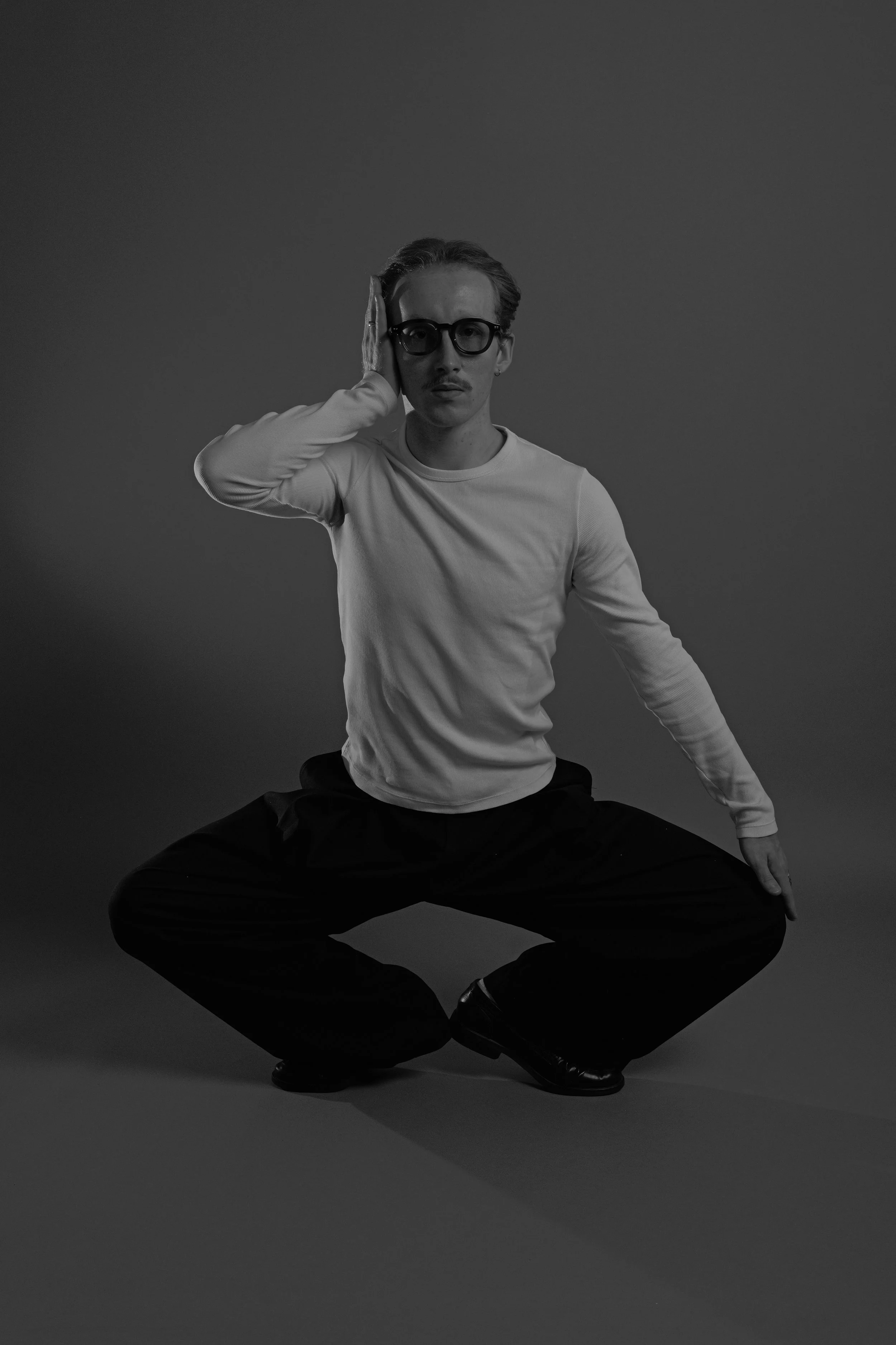 Black and white photo of a man with glasses, sitting in a yoga pose with legs crossed, one hand on his head, against a plain background.