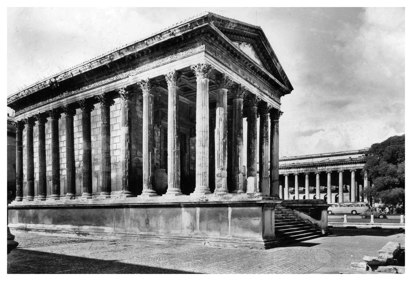 Historical building with classical Greek architectural style, featuring tall columns with ornate capitals, steps leading up to the entrance, and a triangular pediment on top. Black and white photo, possibly an ancient monument or temple.