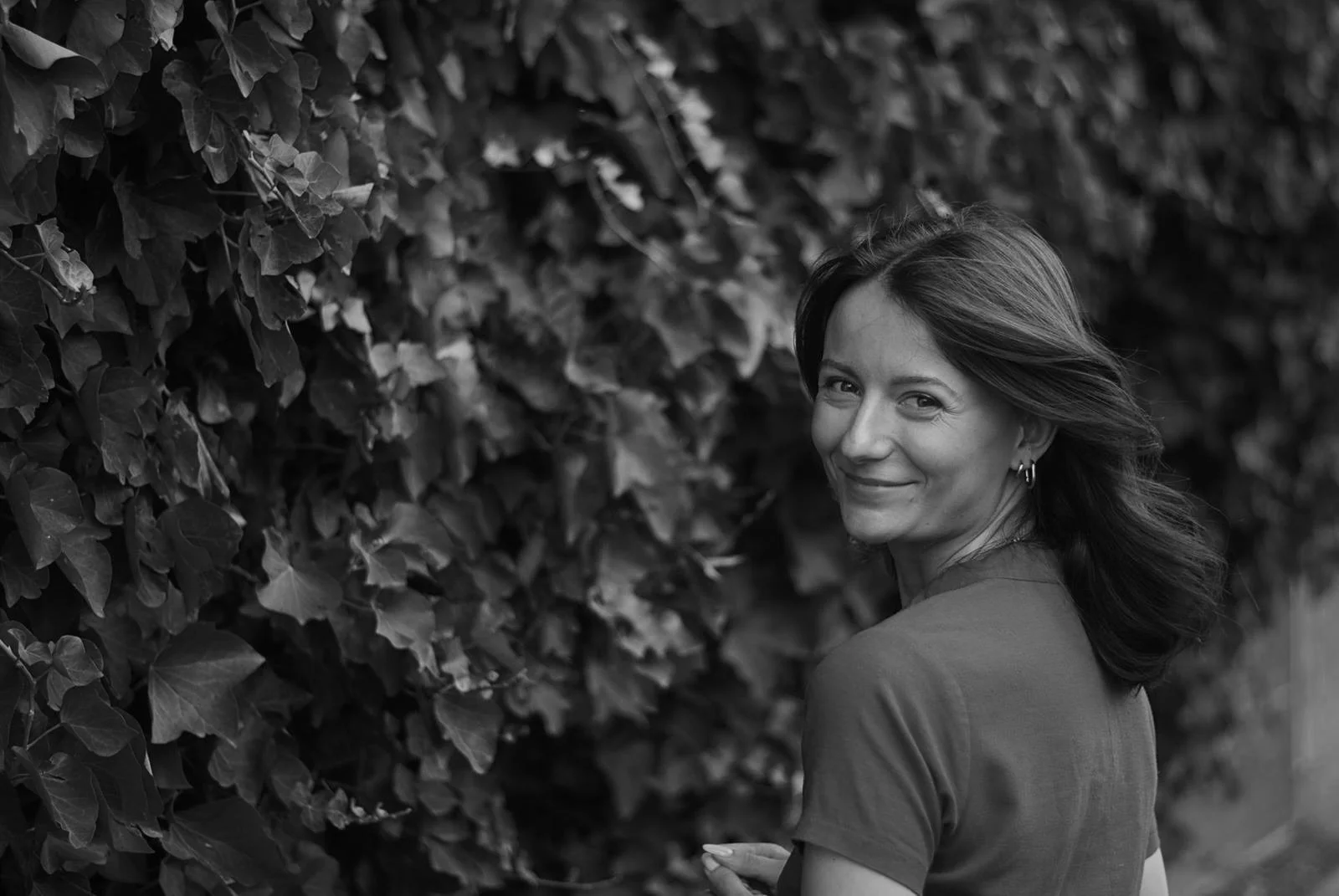 A woman with shoulder-length hair, smiling and looking over her shoulder, standing in front of a wall covered with leaves.