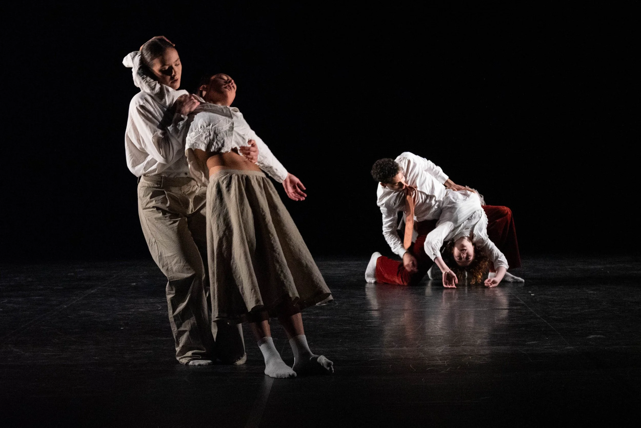 The image shows four dancers on a dark stage, engaging in a dramatic dance performance. Two dancers are standing while two are on the floor, with one leaning on their hands and knees, and the other lying with their head near the floor. They are dressed in neutral-colored costumes.